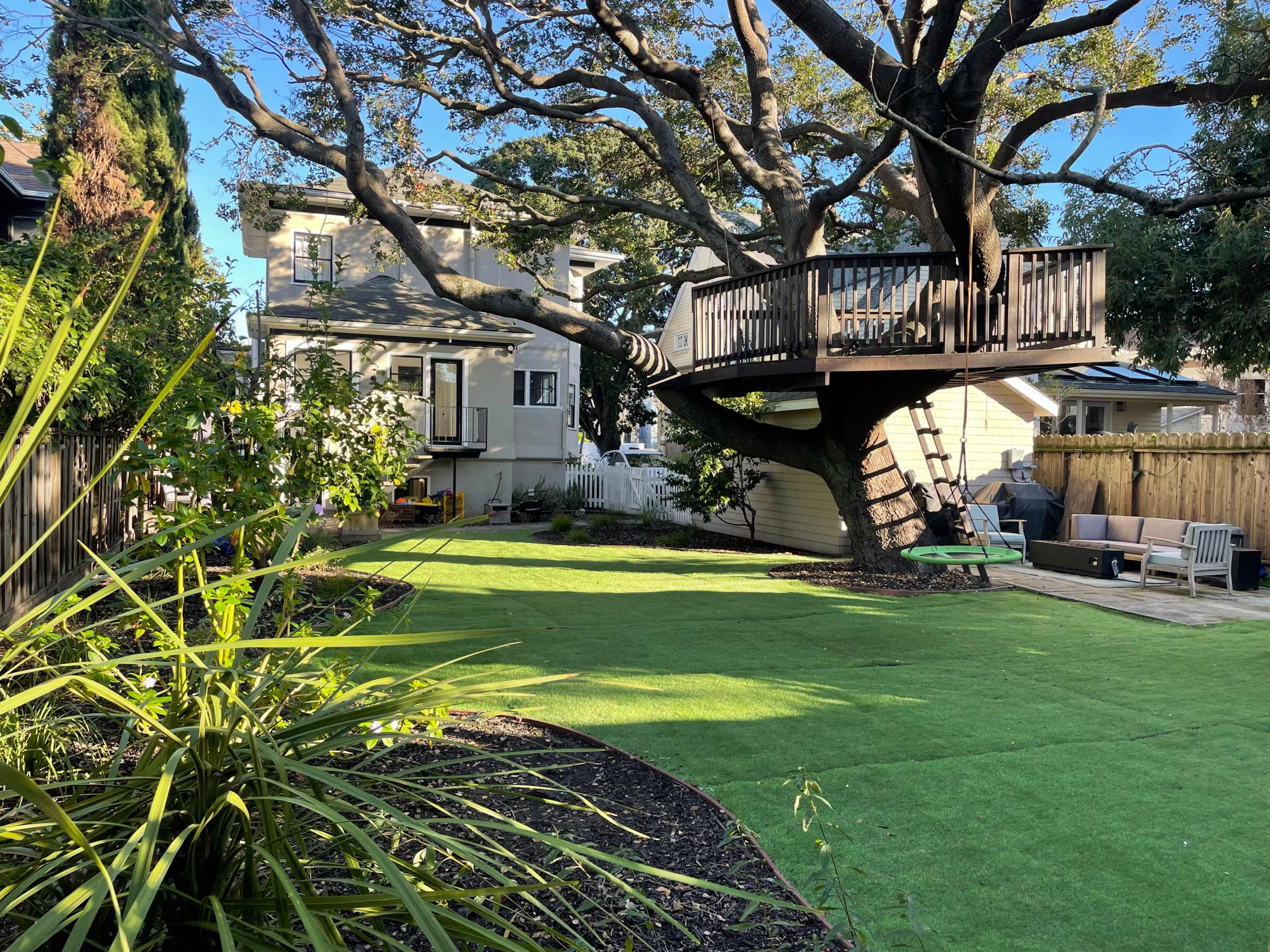 The image shows a backyard with a treehouse built into a large tree, surrounded by green lawn and various plants, with a house in the background.