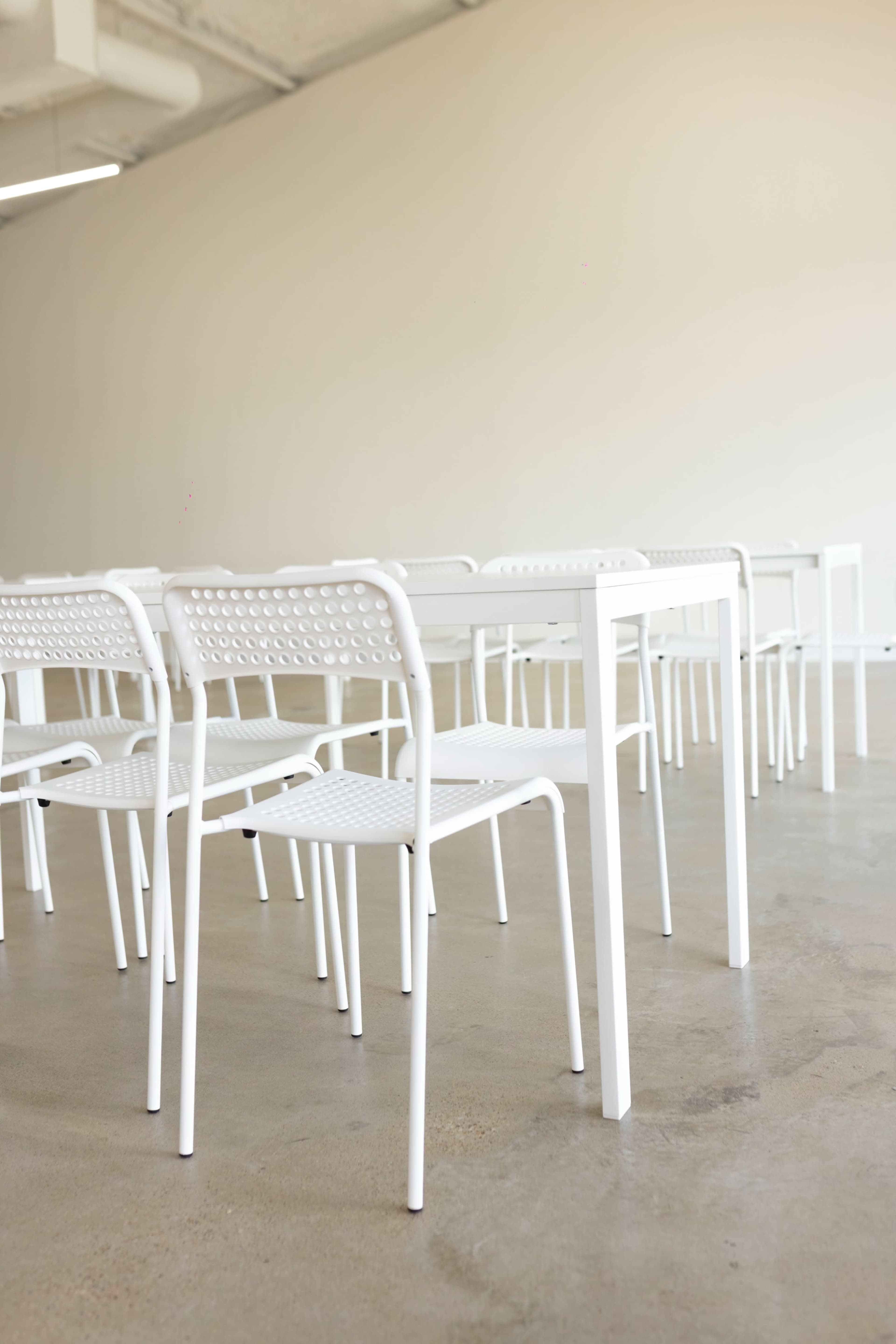 A row of white chairs is arranged in a minimalist indoor space with a concrete floor and light-colored walls.