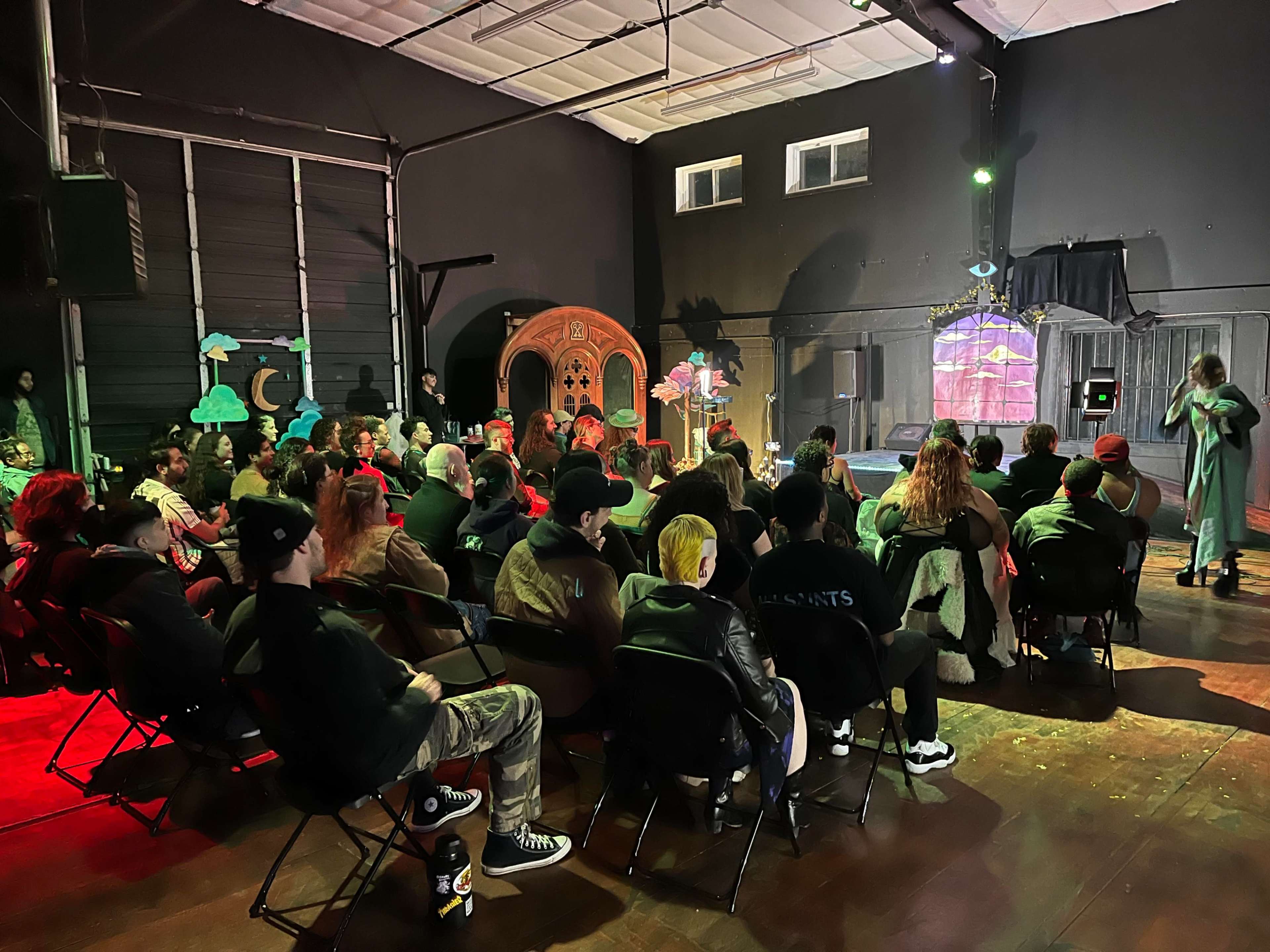 A large audience sits in chairs facing a performer on stage in a dimly lit venue decorated with colorful props.