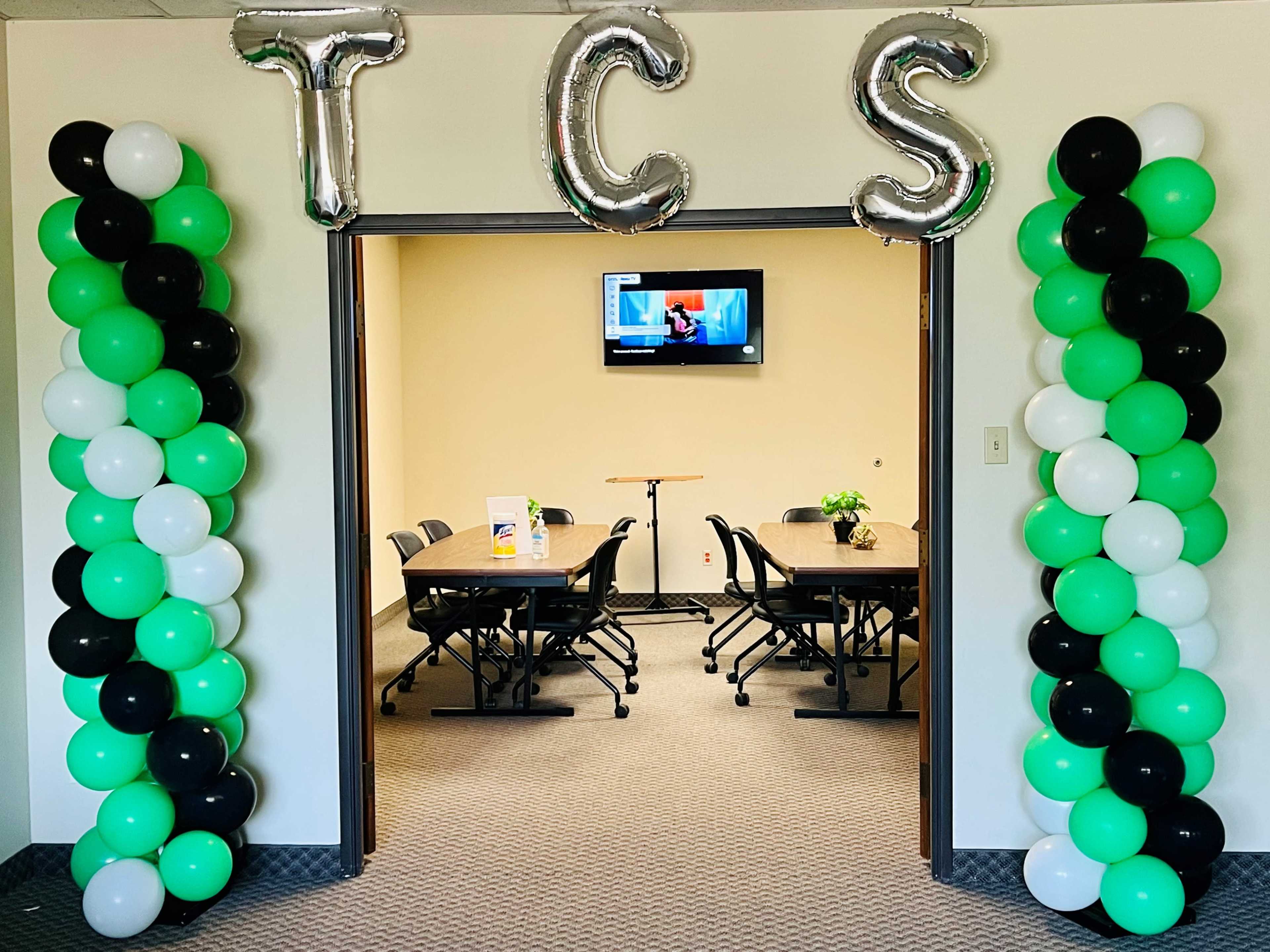 The image shows a doorway with green, black, and white balloon columns on either side, leading into a conference room featuring a table and chairs.