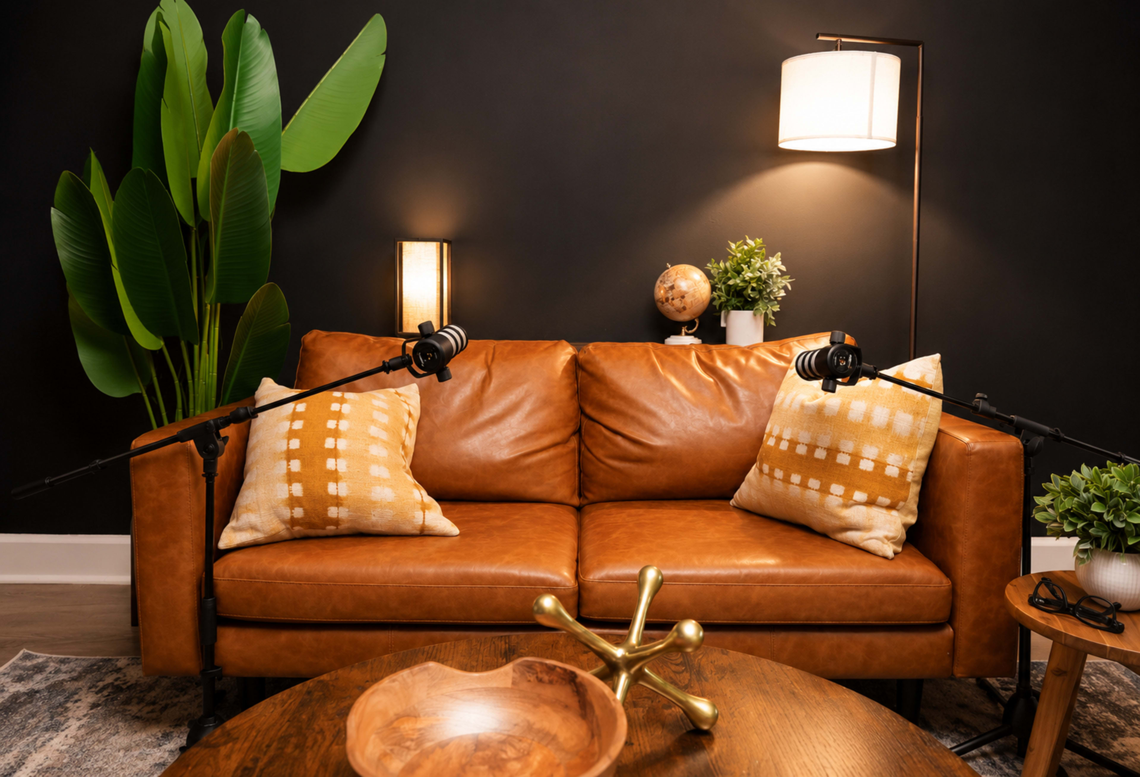 The image shows a brown leather sofa with orange and cream pillows, positioned in a dimly lit room featuring a floor lamp, a globe, and a large plant.