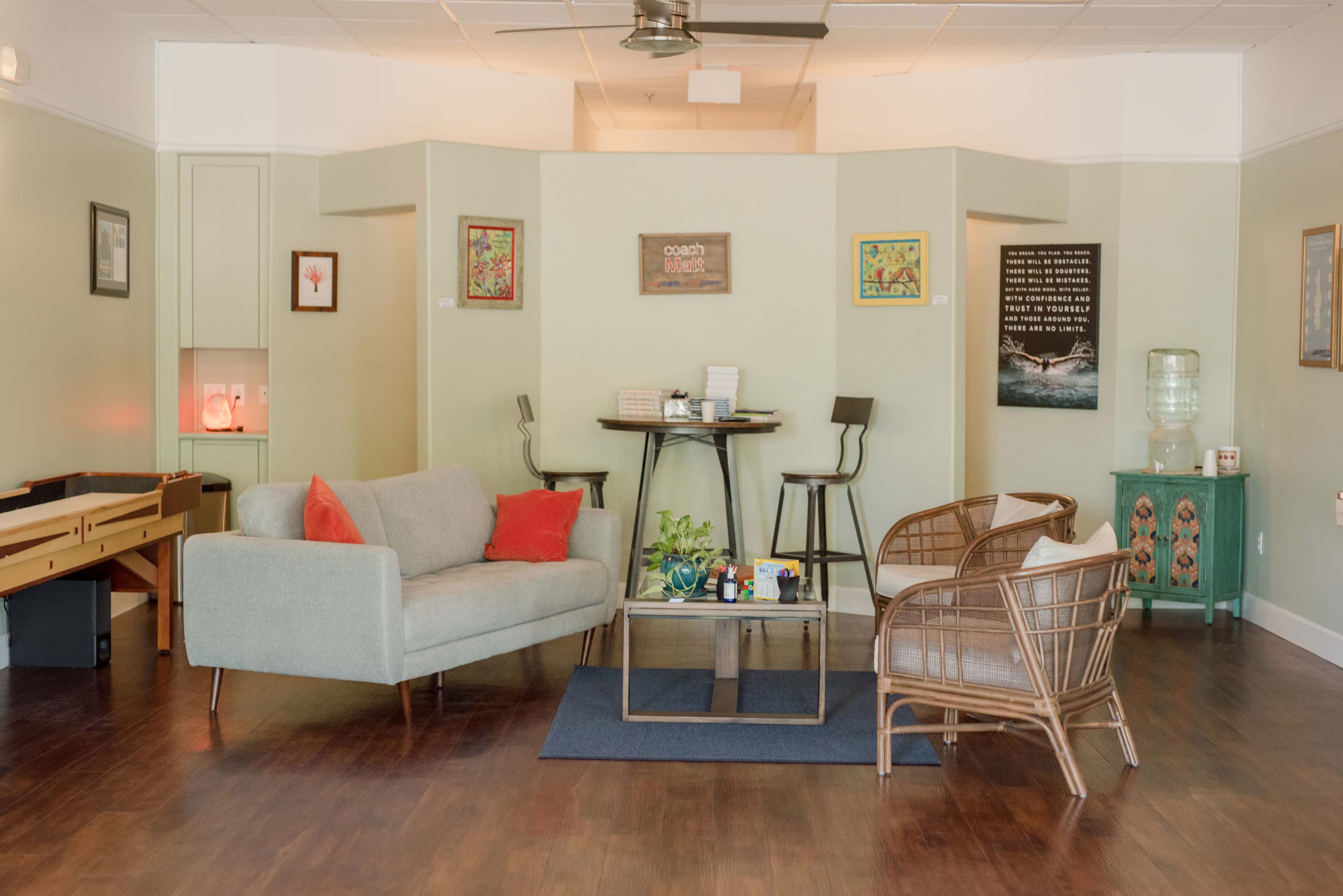A modern waiting area with a grey sofa, two rattan chairs, a glass coffee table, and decorative wall art.
