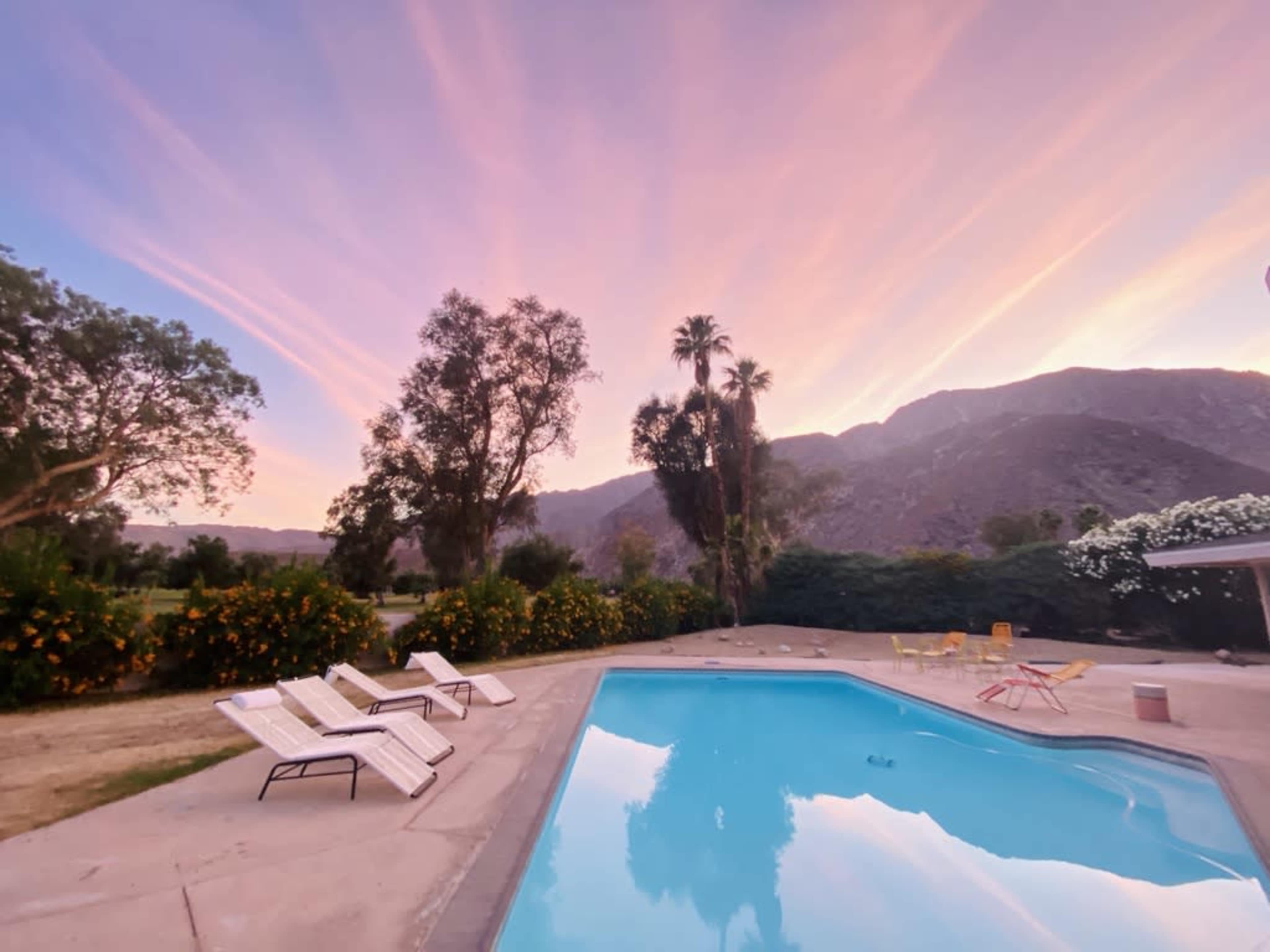 The scene shows a swimming pool surrounded by lounge chairs, with trees and mountains in the background under a colorful sky at sunset.