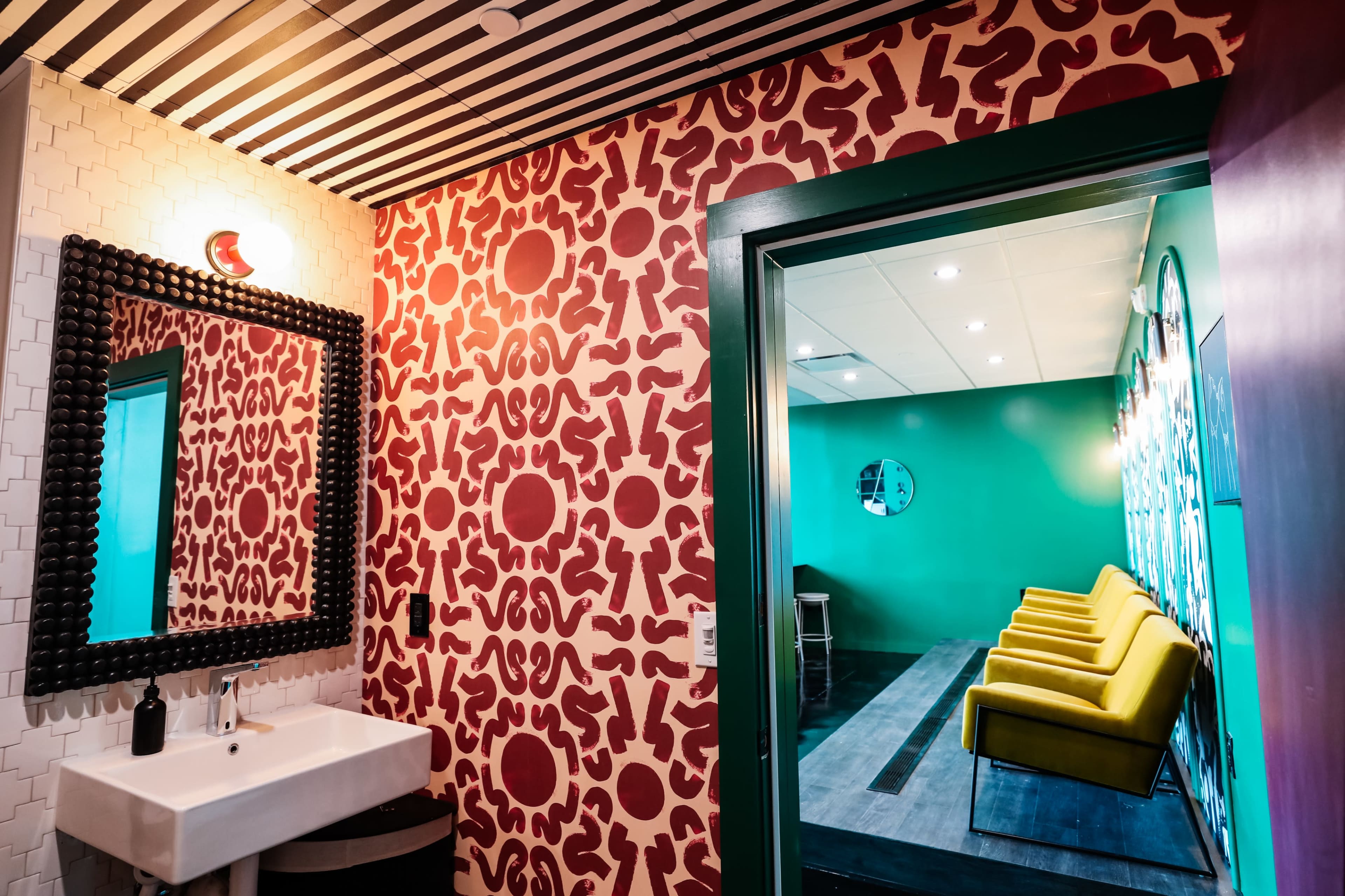 A brightly colored restroom features a patterned red wall and a large mirror, with an open door leading to a room with yellow chairs against a green wall.
