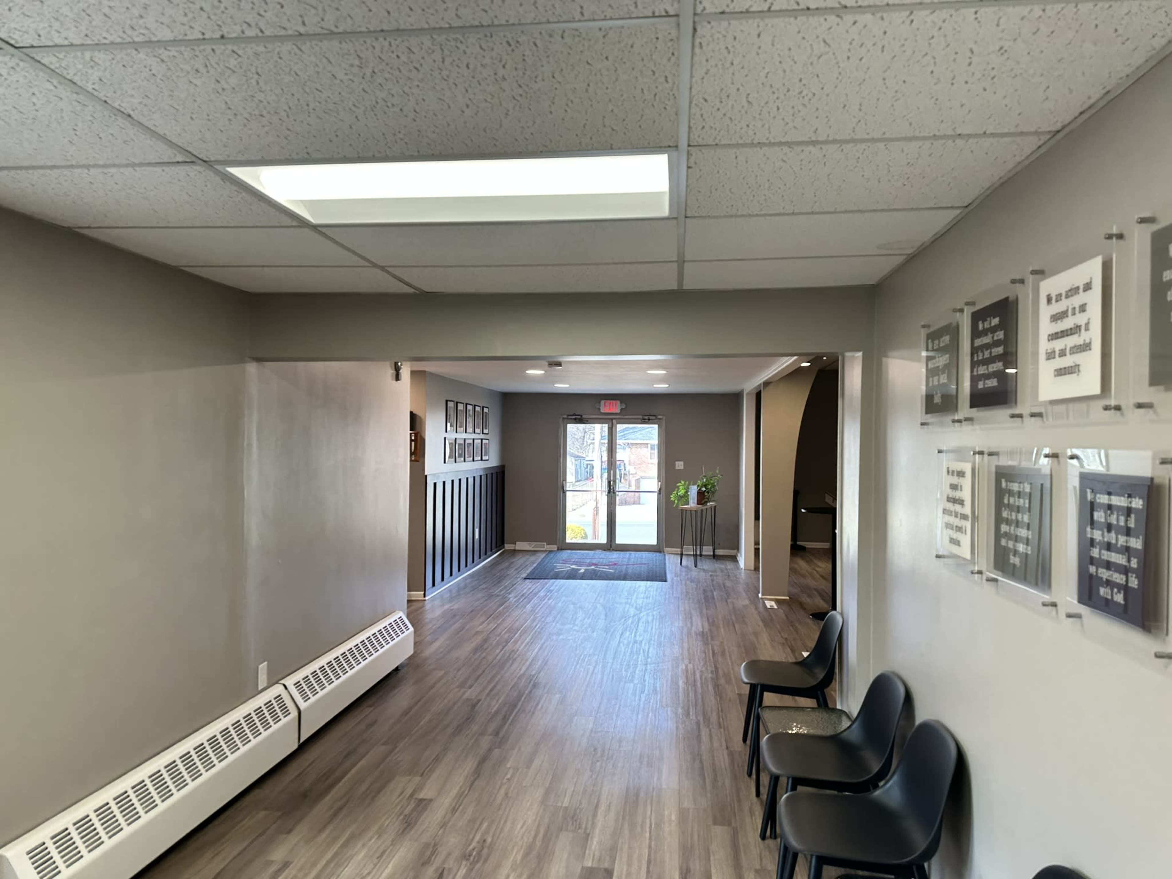 A hallway with a clean floor, leading to a door at the end, flanked by black chairs and framed signs on the walls.