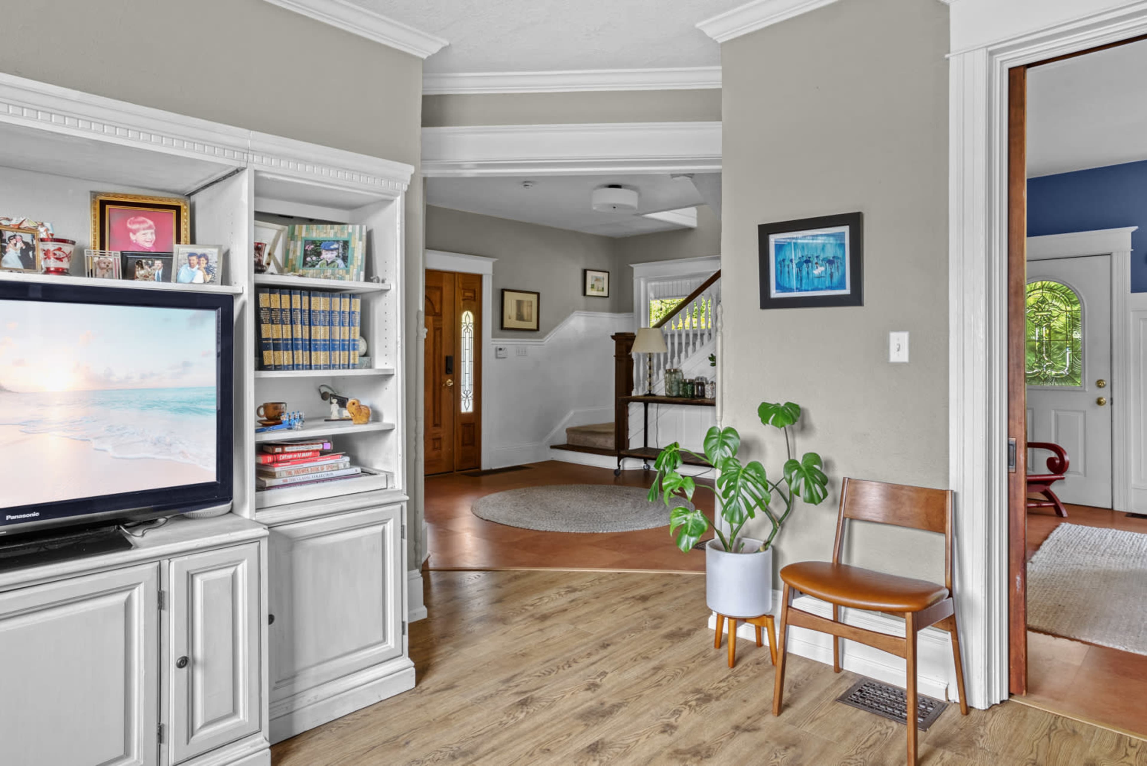 The image shows a light-colored living room with built-in shelves, a television, and a chair near a green plant, leading towards a hallway and entrance area.