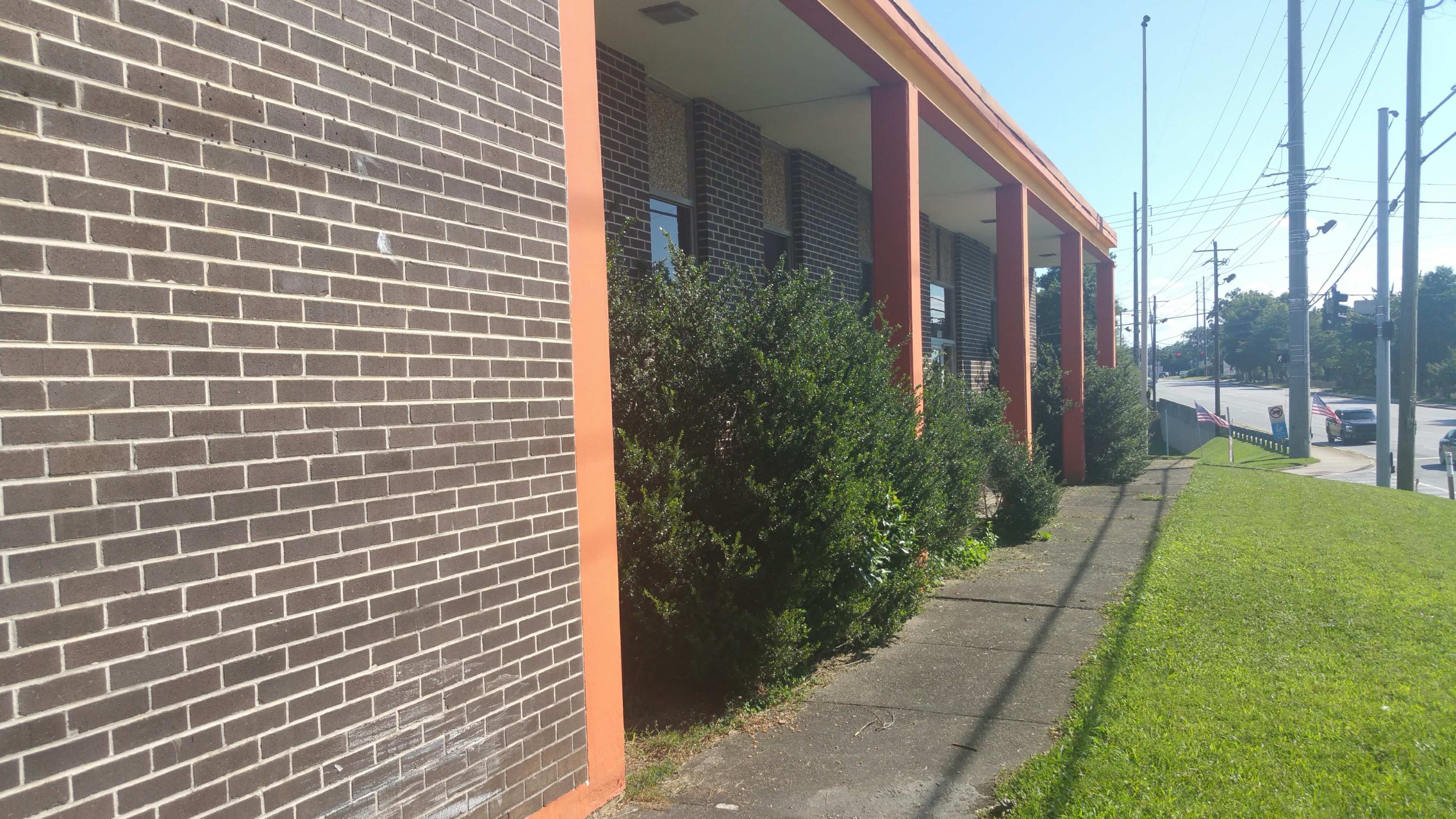 The image shows a brick building with orange accents, bordered by shrubs on a concrete path next to a grassy area.