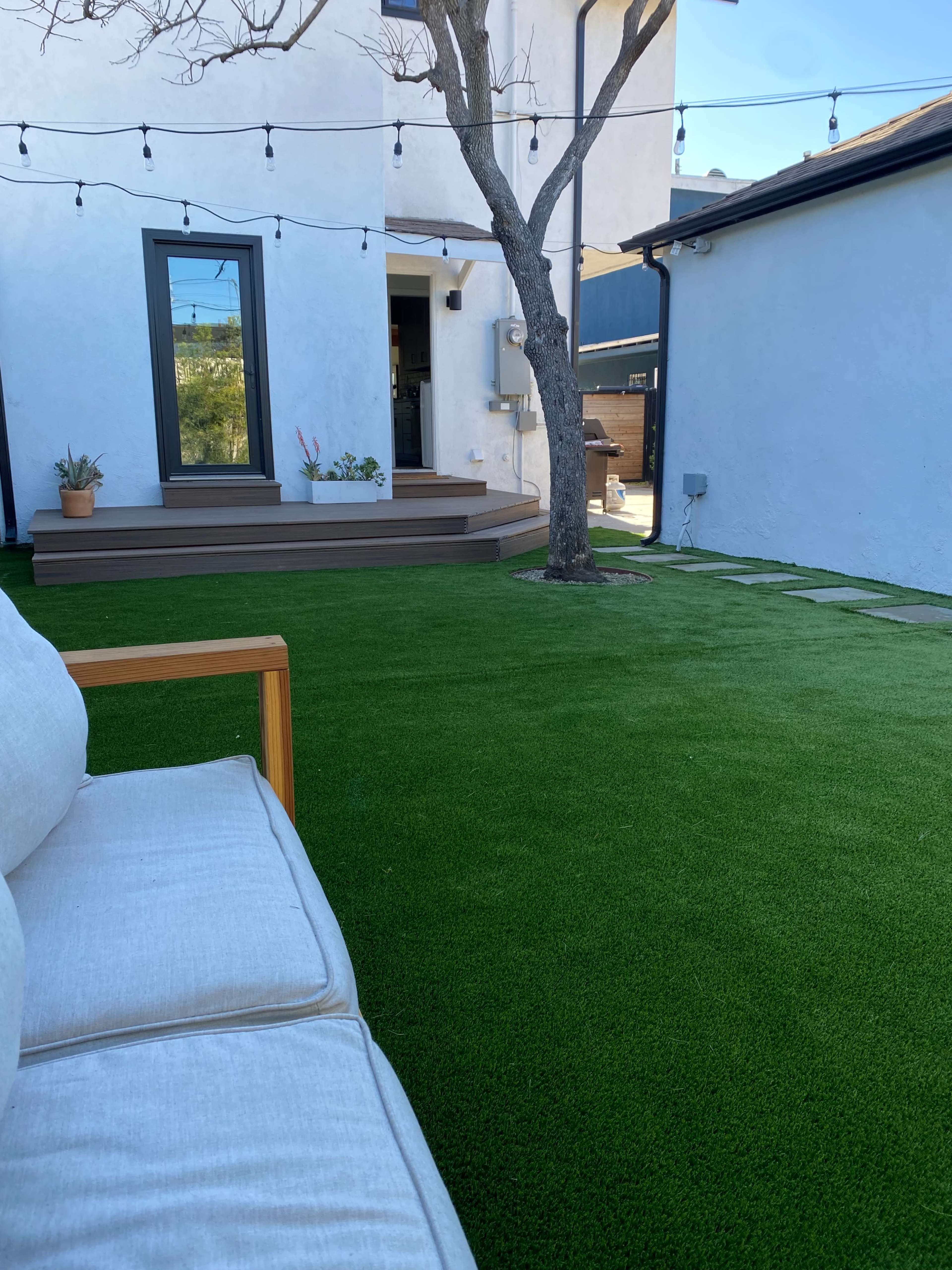 The image shows a backyard with artificial turf, a wooden deck, a tree, and a view of a modern white building.