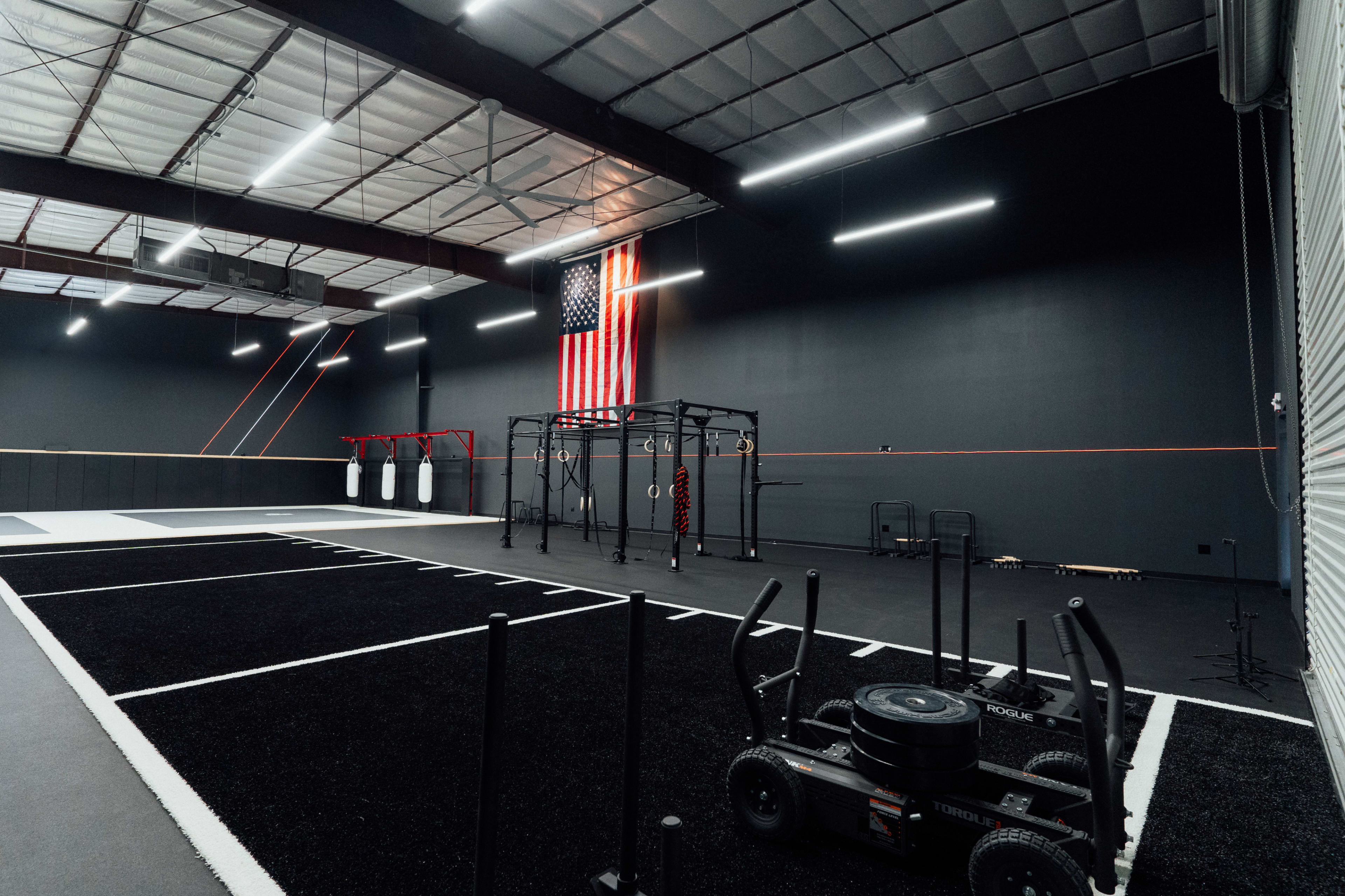 An indoor gym facility with a dark gray color scheme, featuring a training area with weightlifting equipment, a flag hanging on the wall, and marked spaces on the floor for various exercises.