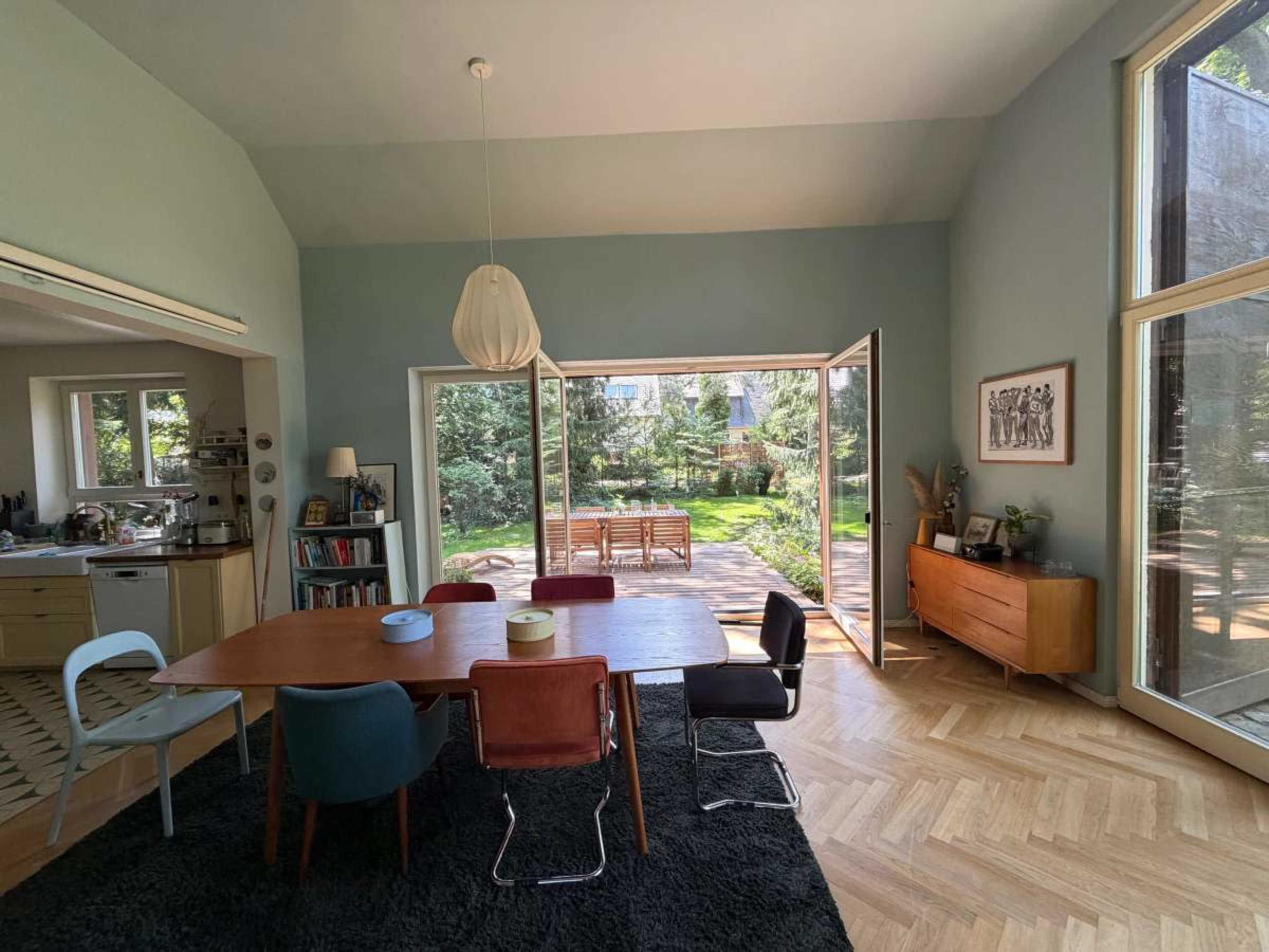 The image shows a modern dining area with a large table surrounded by chairs, open to a backyard garden through sliding glass doors.