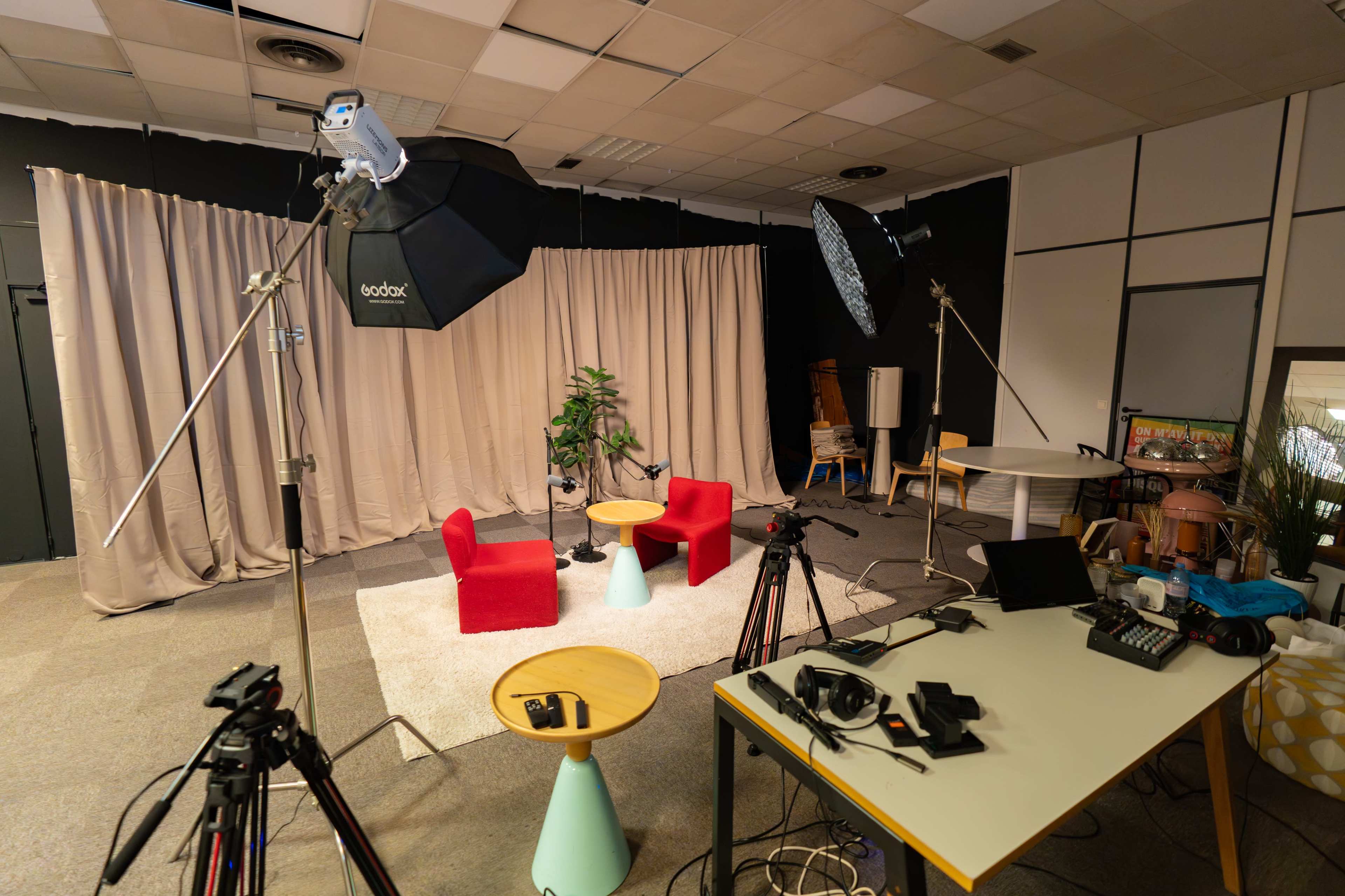 The image shows a studio setup with two red chairs, a small table, lighting equipment, and cameras arranged around a backdrop of beige curtains.