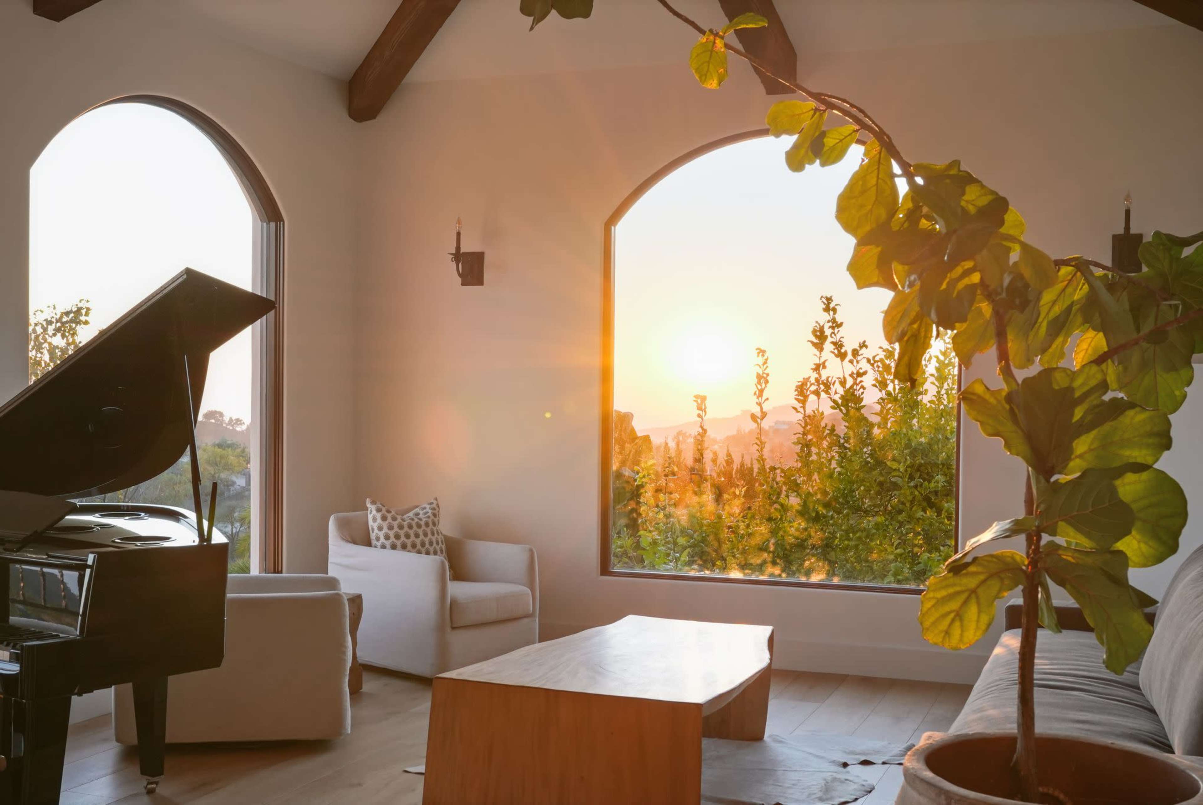 A sunlit living room features a grand piano, a wooden coffee table, and large arched windows showcasing a sunset over greenery.