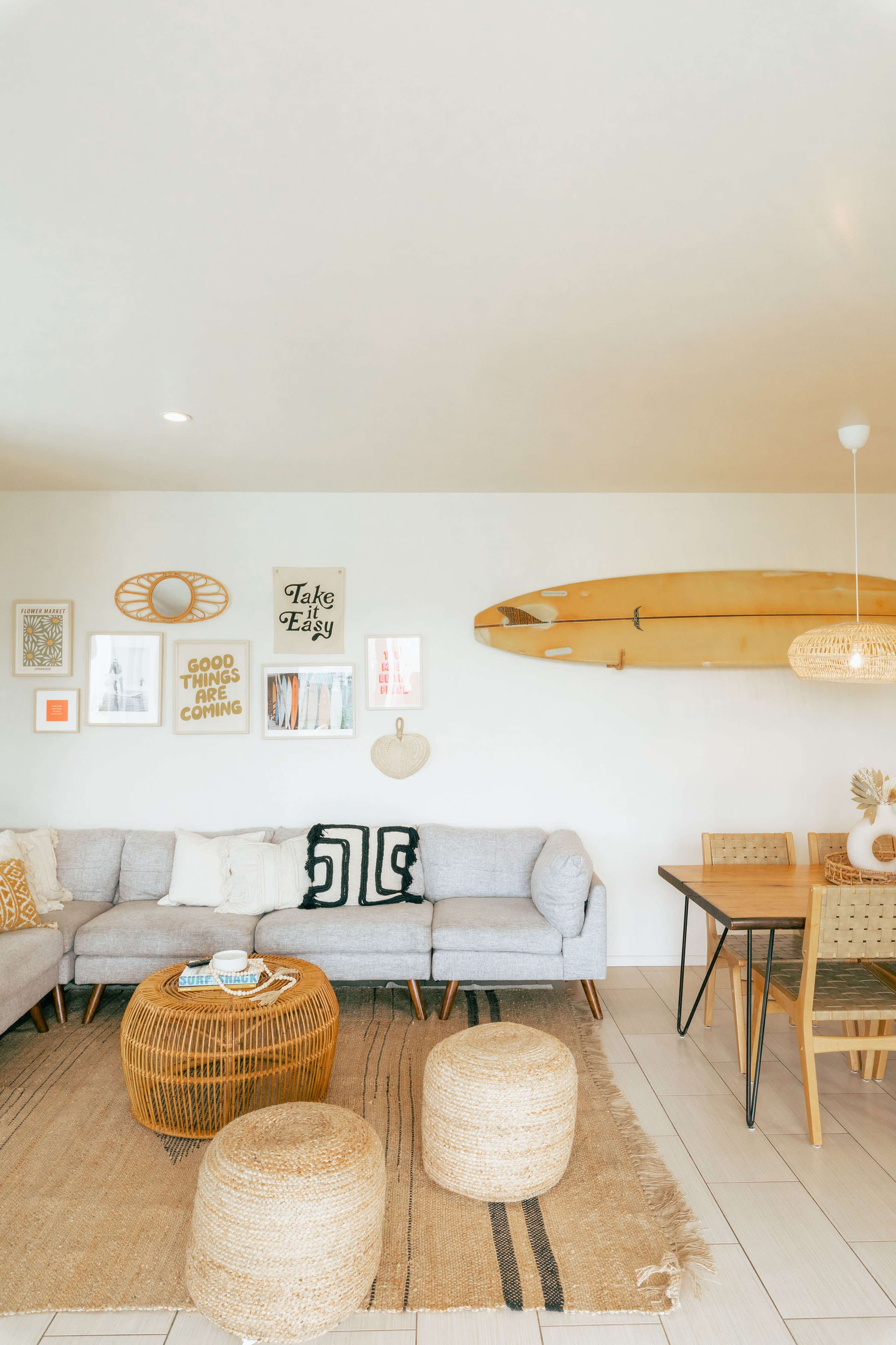 A cozy living room features a light gray sofa, wall art including framed quotes, a wooden surfboard mounted above, and a dining area with a round table and woven stools.