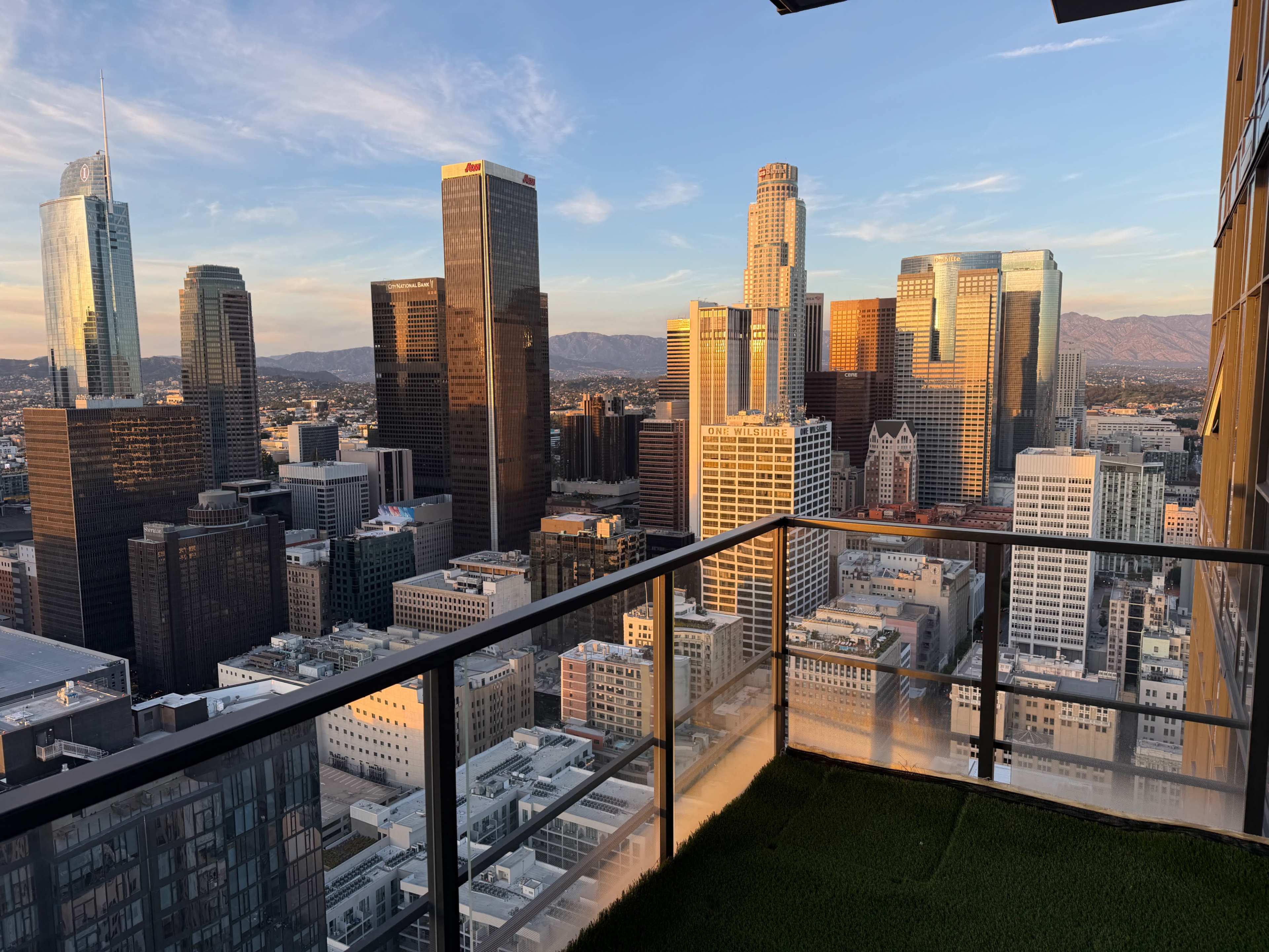 The image shows a panoramic view of a city skyline at sunset, with tall buildings and a balcony in the foreground.