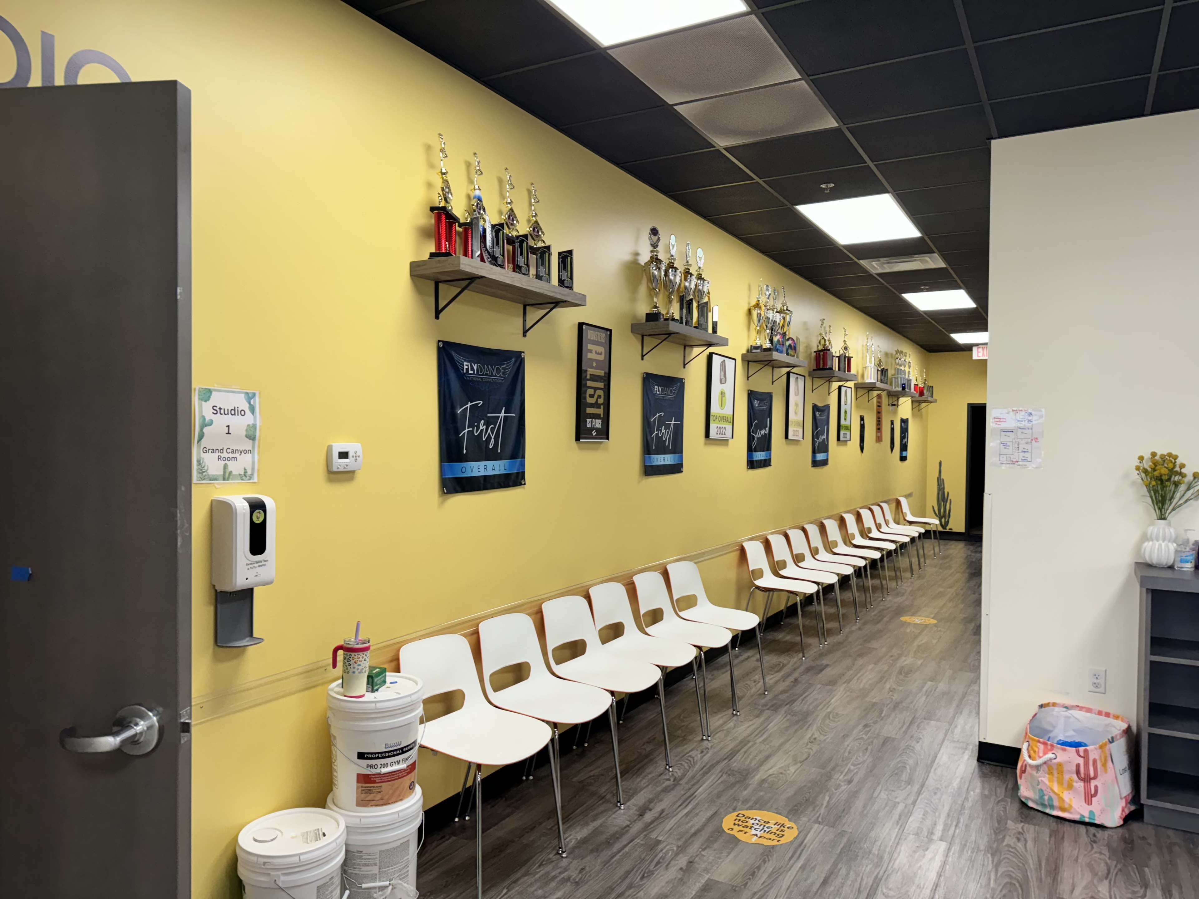 A long hallway with yellow walls, lined with chairs on one side and shelves displaying trophies and framed posters on the other.