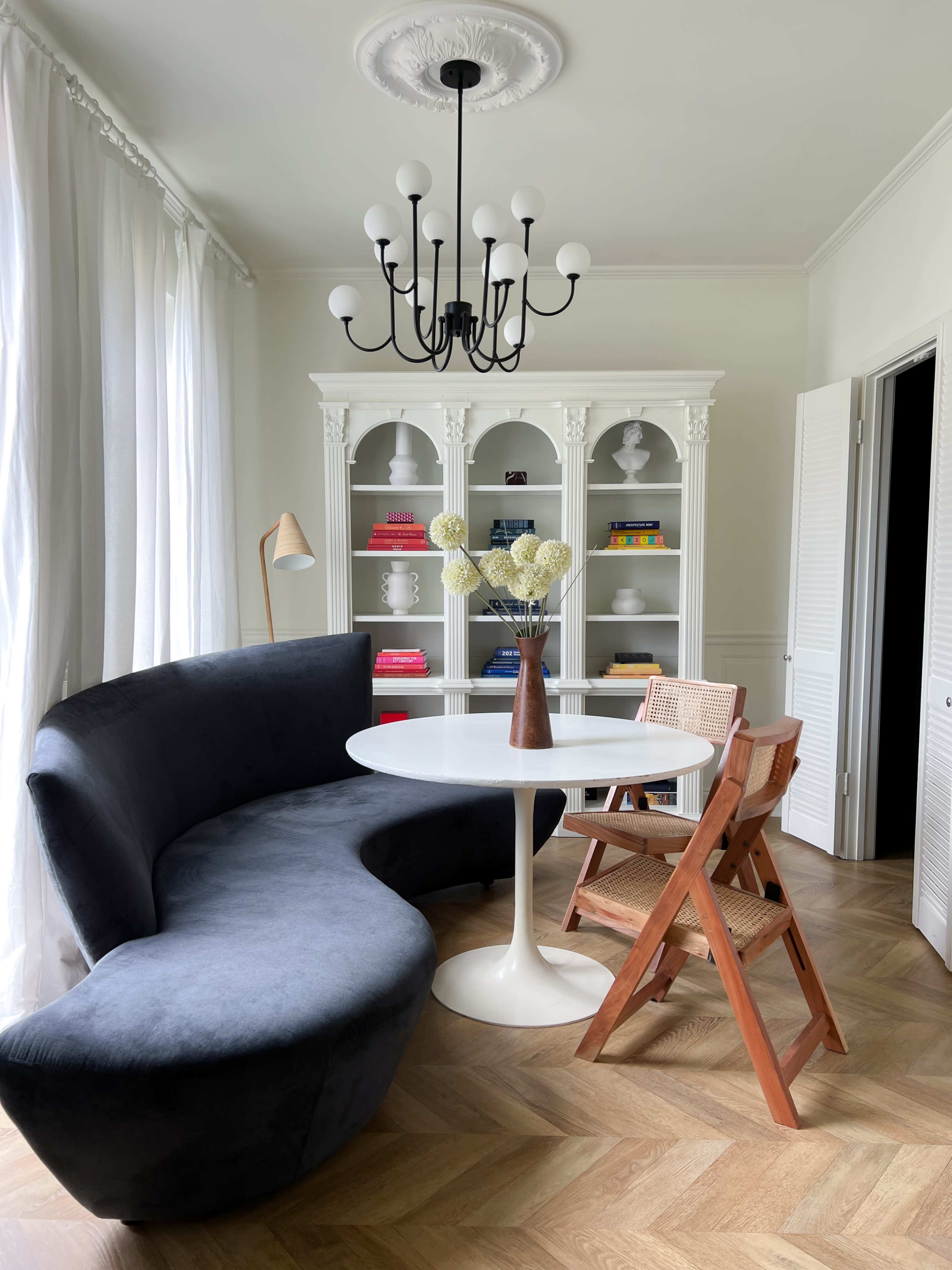 The image shows a modern, curved gray sofa facing a round white dining table surrounded by wooden chairs in a well-lit room with a chandelier and bookshelves.