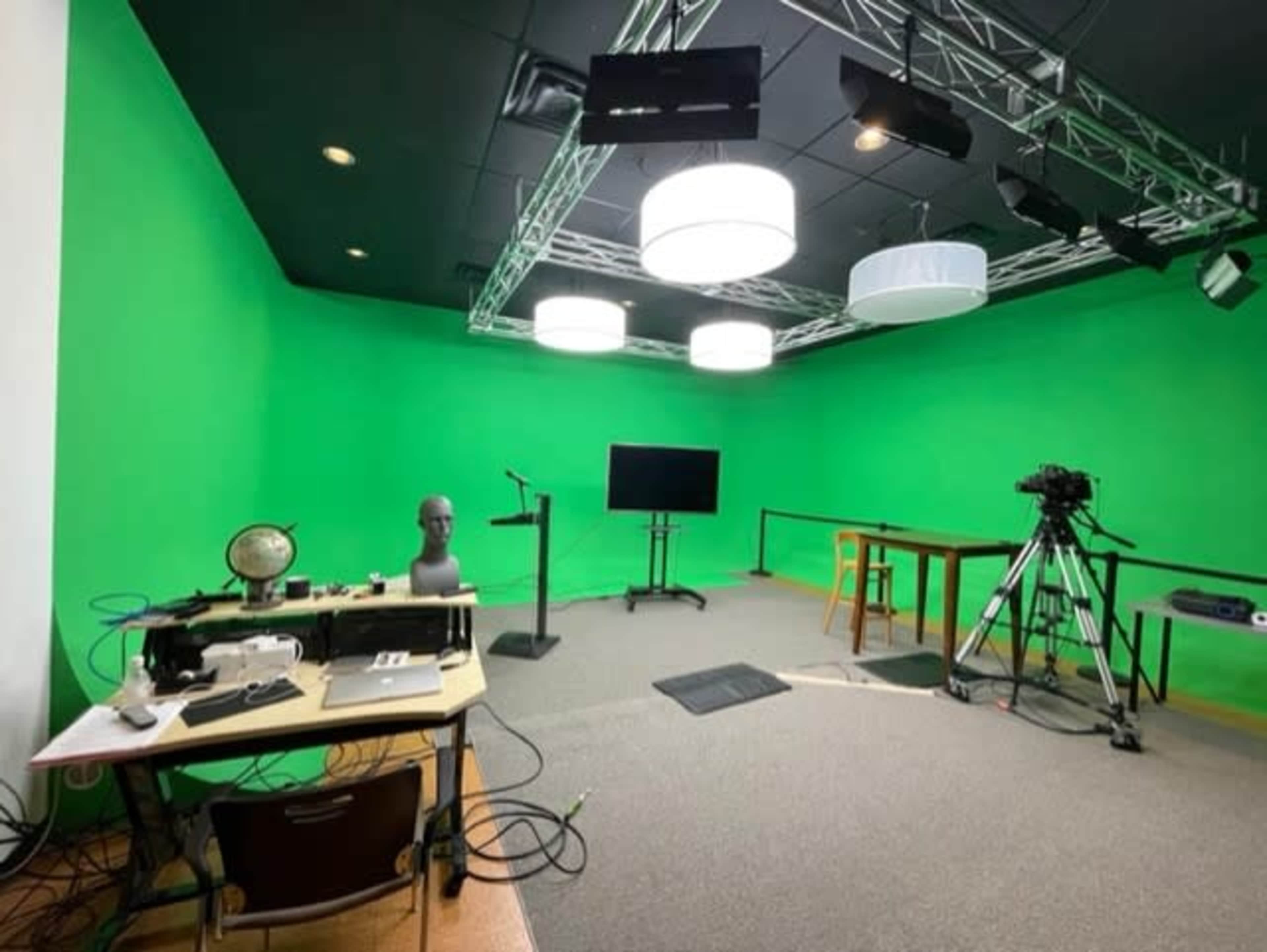 A green-screen studio with various lighting fixtures, a camera setup, and a desk with equipment.