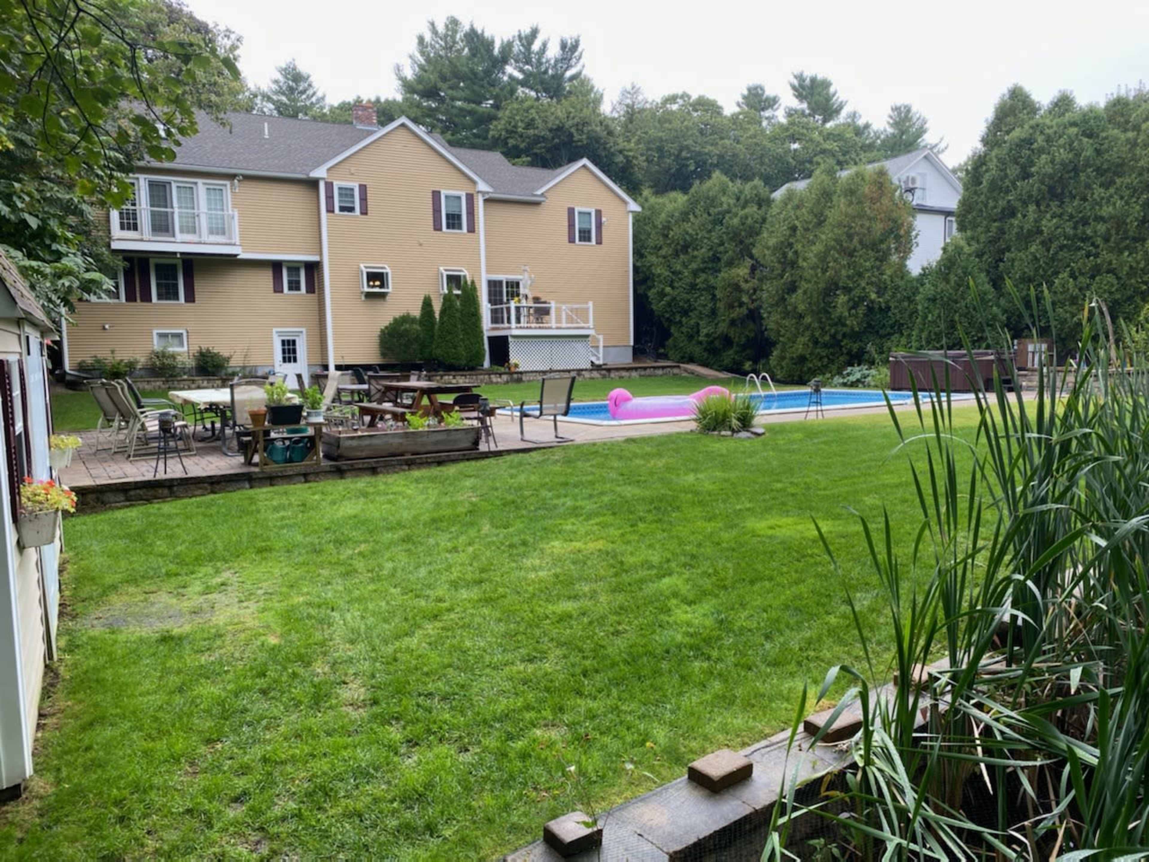 A backyard with a pool, lawn chairs, and a large green lawn in front of a two-story yellow house.