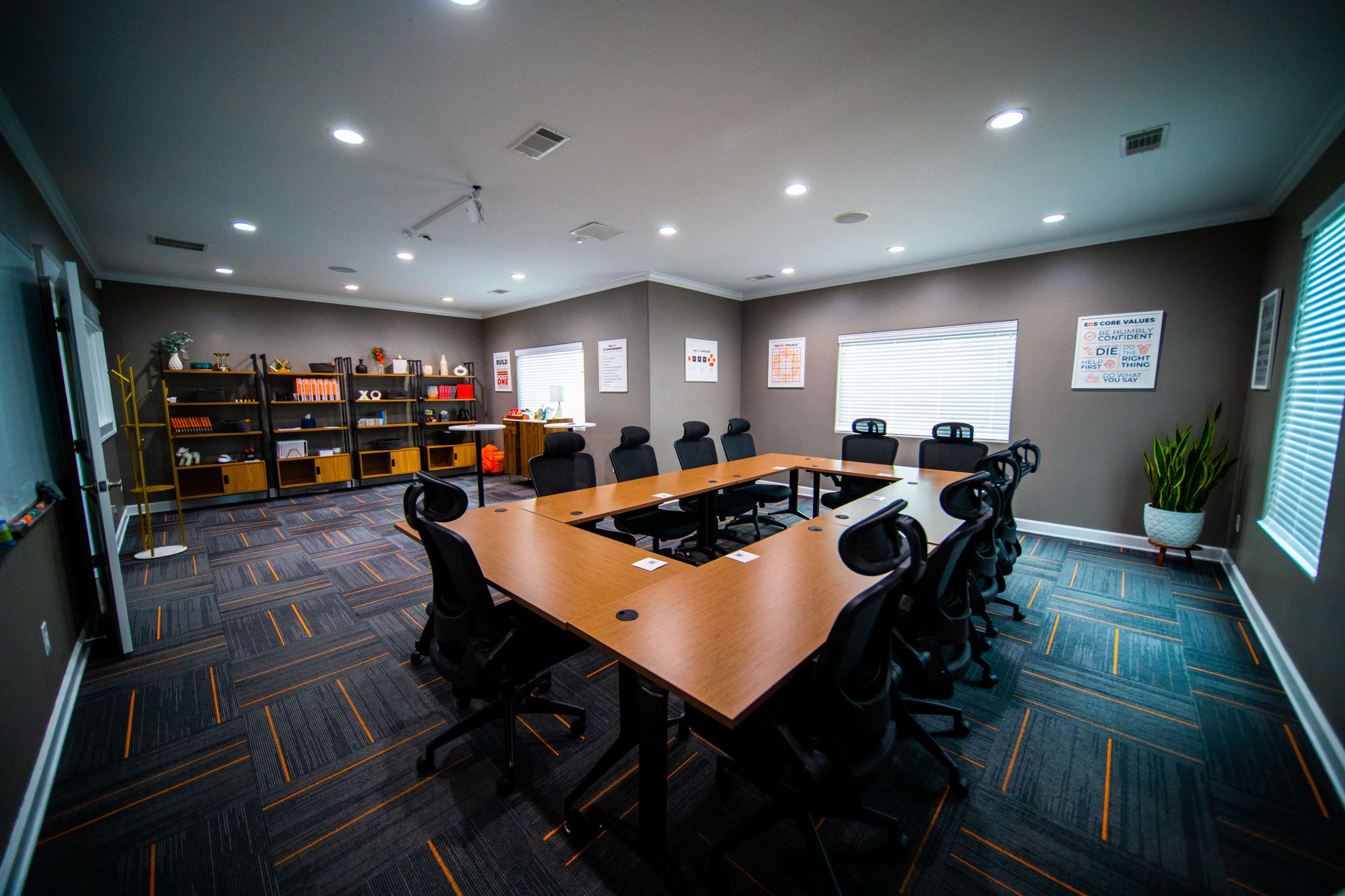 A modern conference room with a large wooden table, black office chairs, and shelves filled with various items.