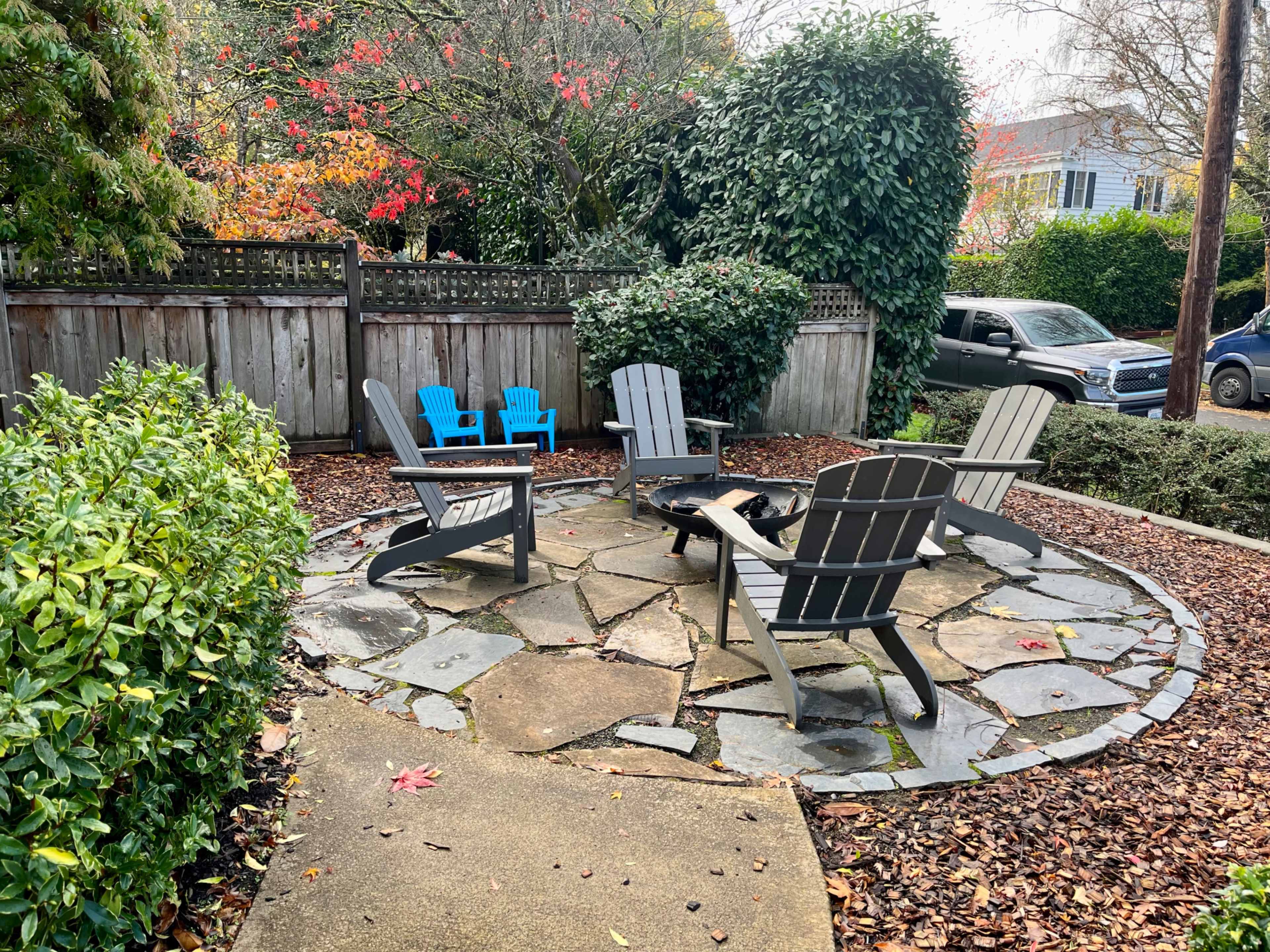 A circular stone patio surrounded by green shrubs, featuring four dark gray Adirondack chairs and two blue chairs.