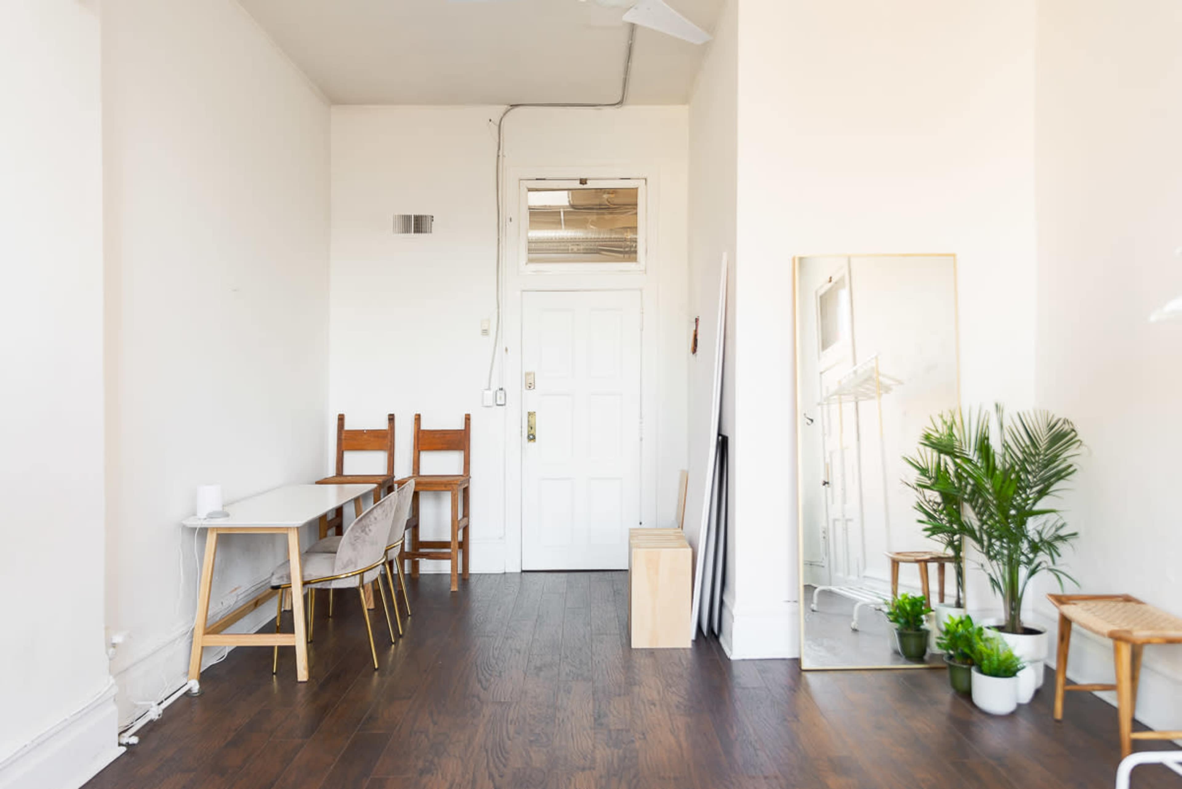 A minimalist room with a wooden floor, a table with chairs, a large mirror, and plants near the corner.