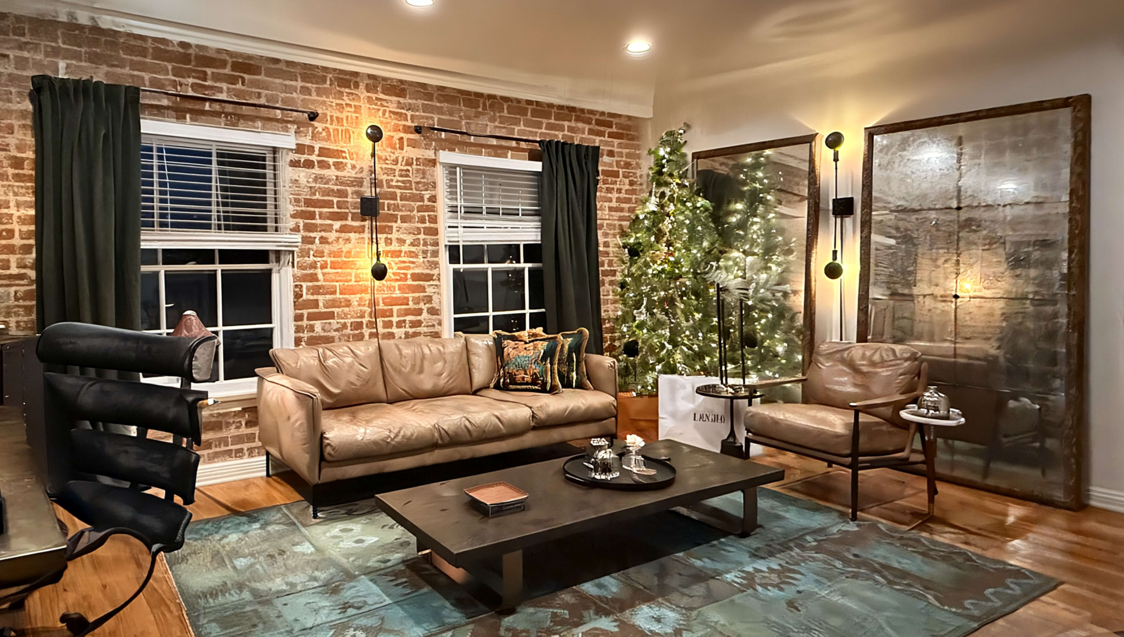 A modern living room features a leather sofa and armchair, a coffee table, a decorative rug, and a Christmas tree beside exposed brick walls and large windows.