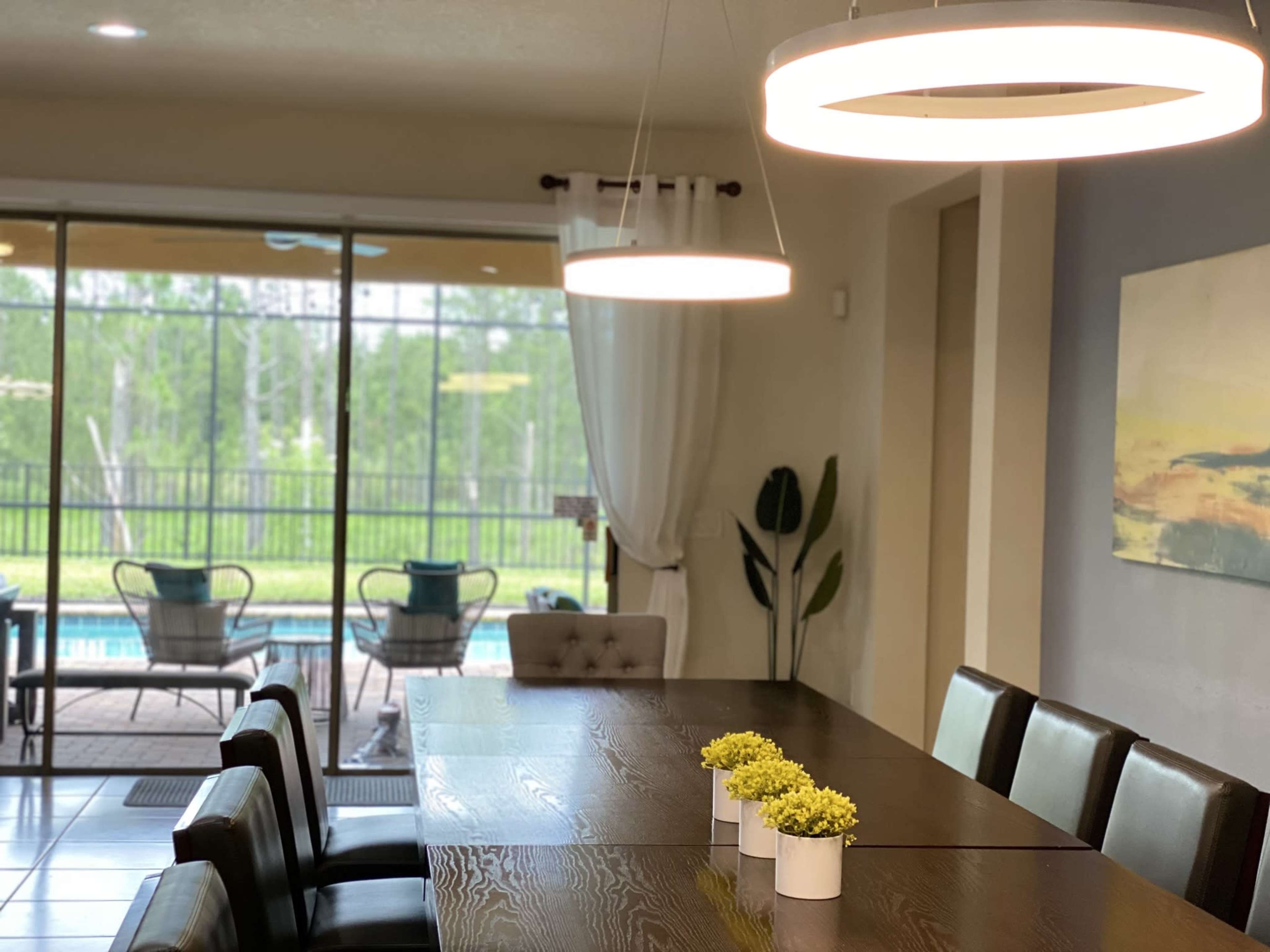 A modern dining area features a long wooden table with black chairs, illuminated by circular pendant lights, and overlooks a backyard with a pool and greenery through large glass doors.