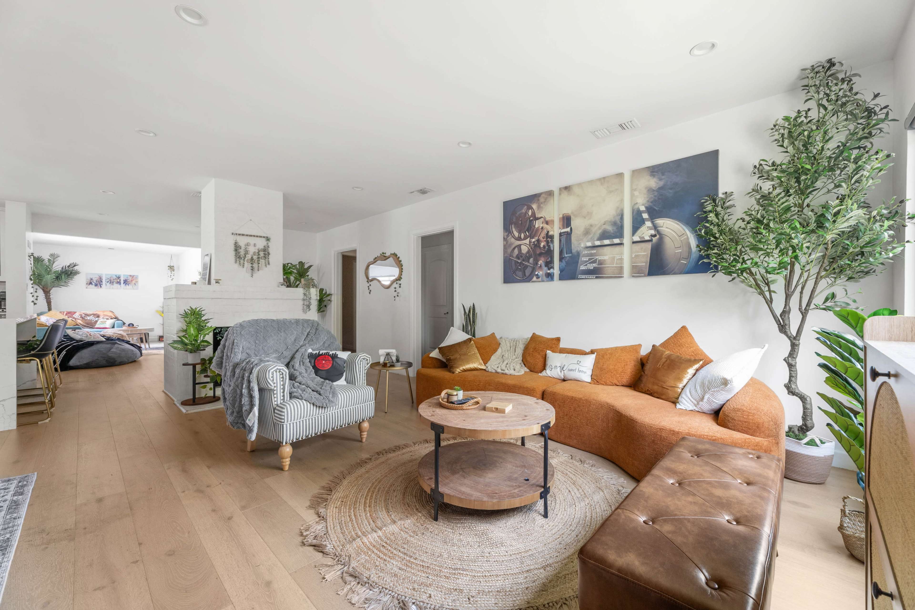 The image shows a bright living room featuring a large, curved brown sofa, a round wooden coffee table, and decorative plants, with an adjoining space that includes a cozy bedroom area.