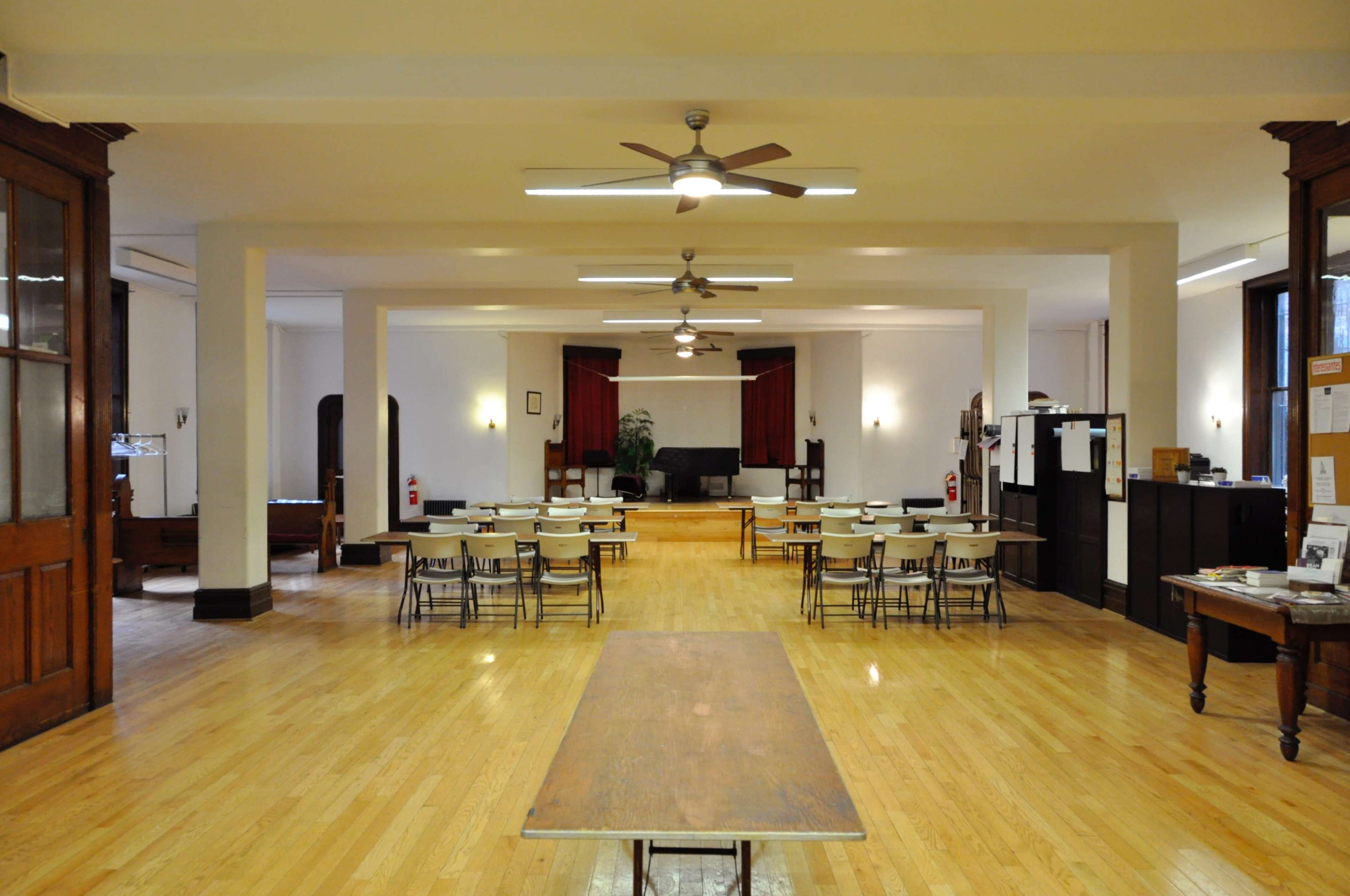 A spacious, empty room with wood flooring, arranged with tables and chairs, and featuring ceiling fans and large windows.