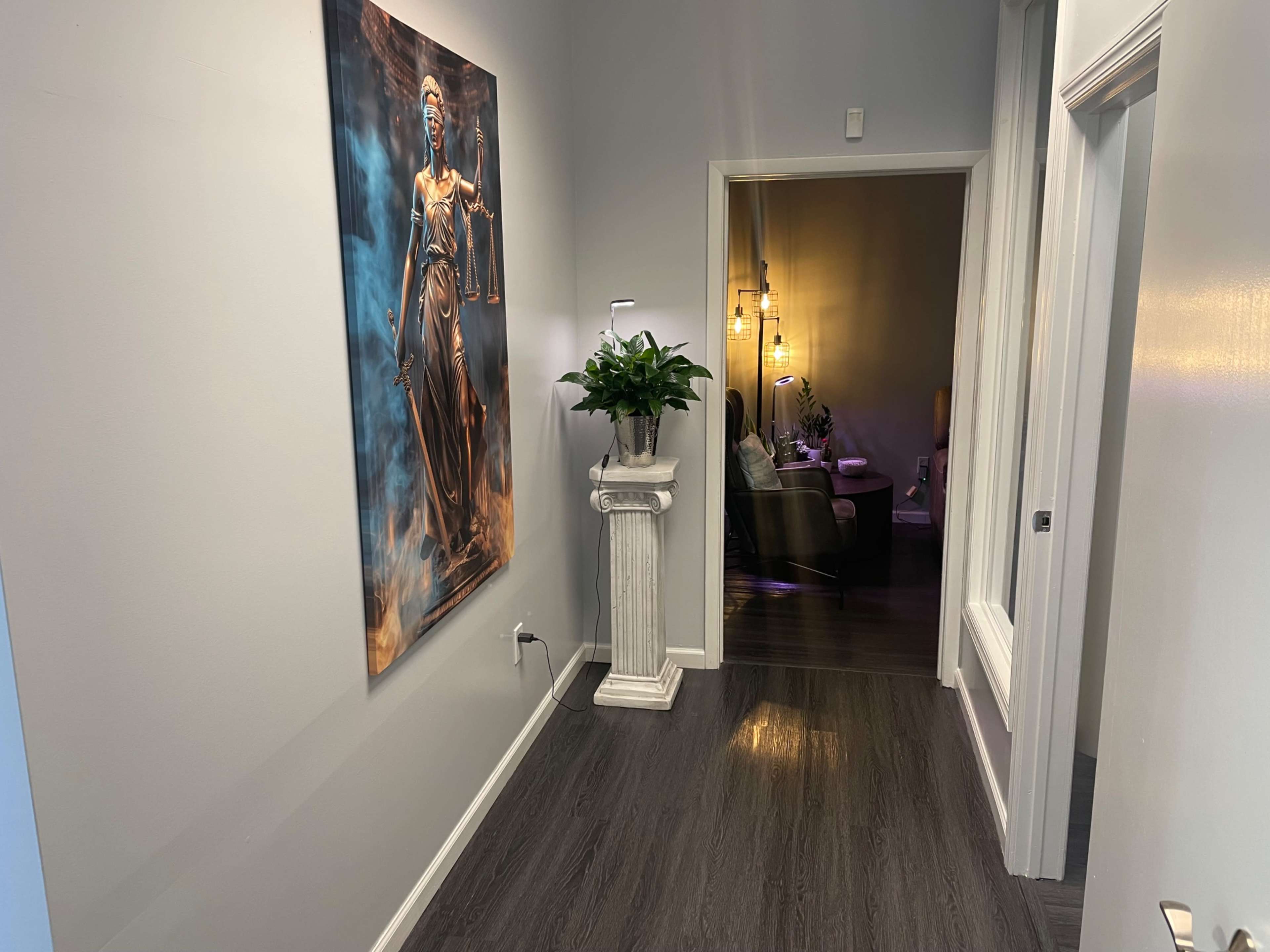 The image shows a hallway with a large painting of a figure holding scales on one wall and a potted plant on a pedestal near the entrance to a living area.