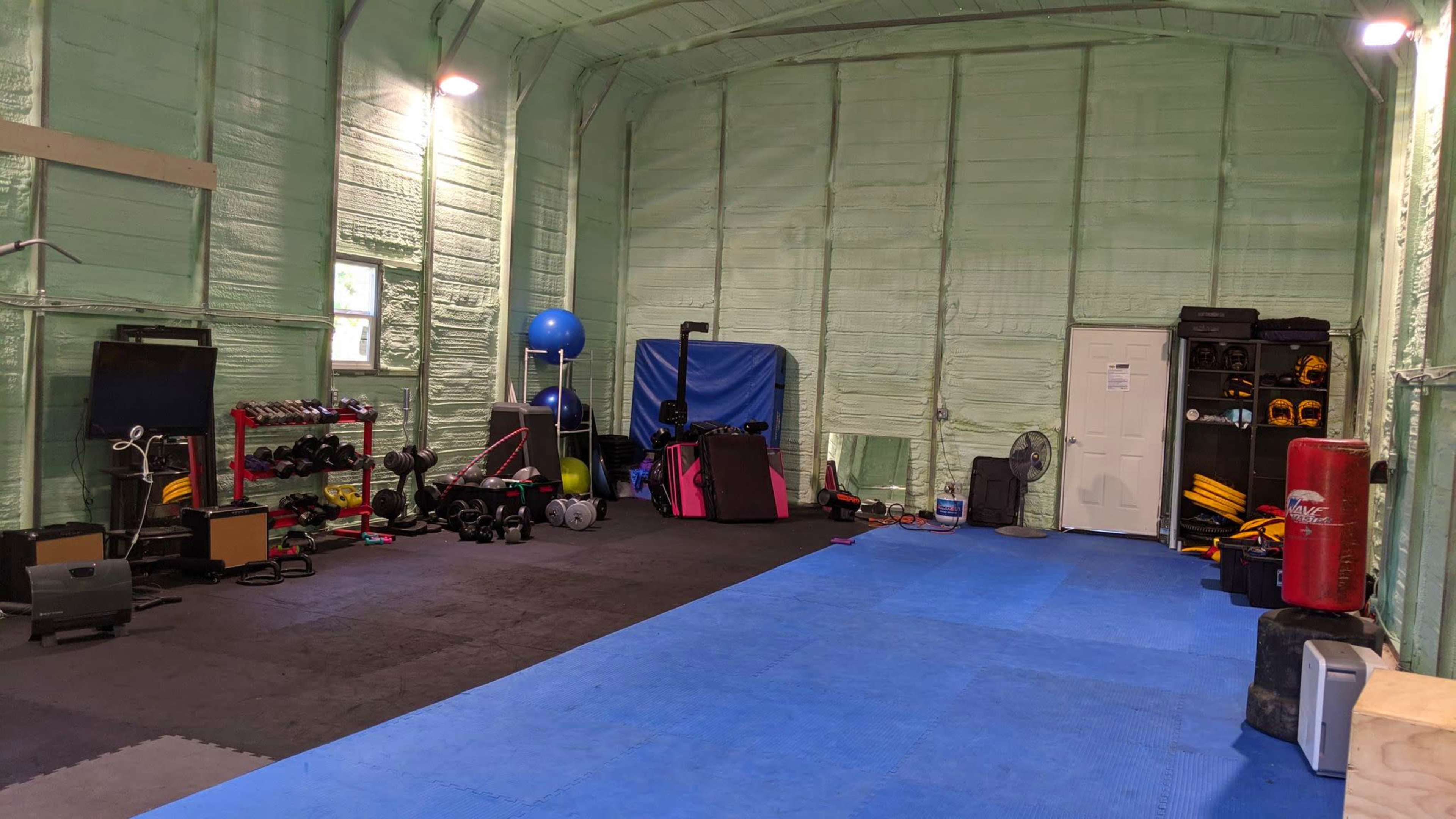 The image shows a gym interior with exercise equipment, including weights, mats, and fitness balls, arranged in a spacious room with green insulated walls.