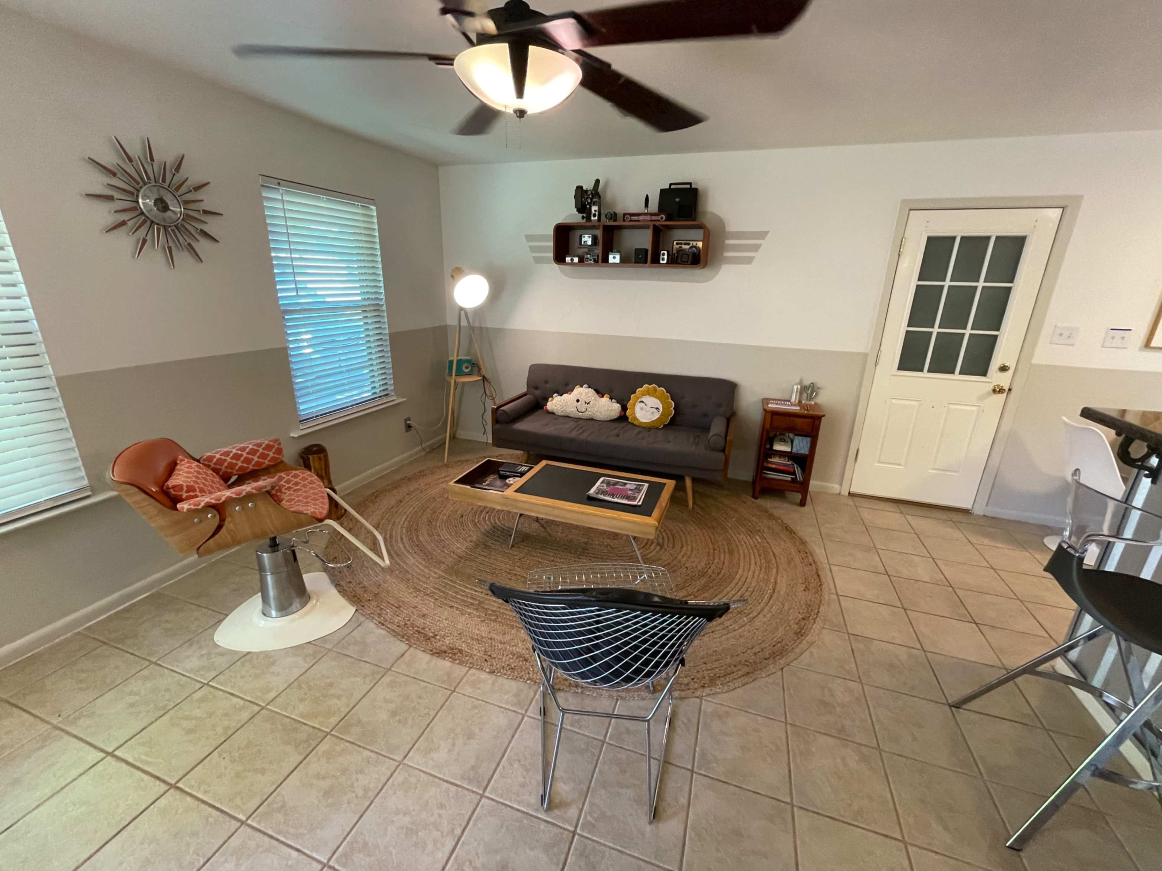 The image shows a living room with a gray sofa, a round rug, a coffee table, and a wooden chair, all arranged around a central space and illuminated by natural light.