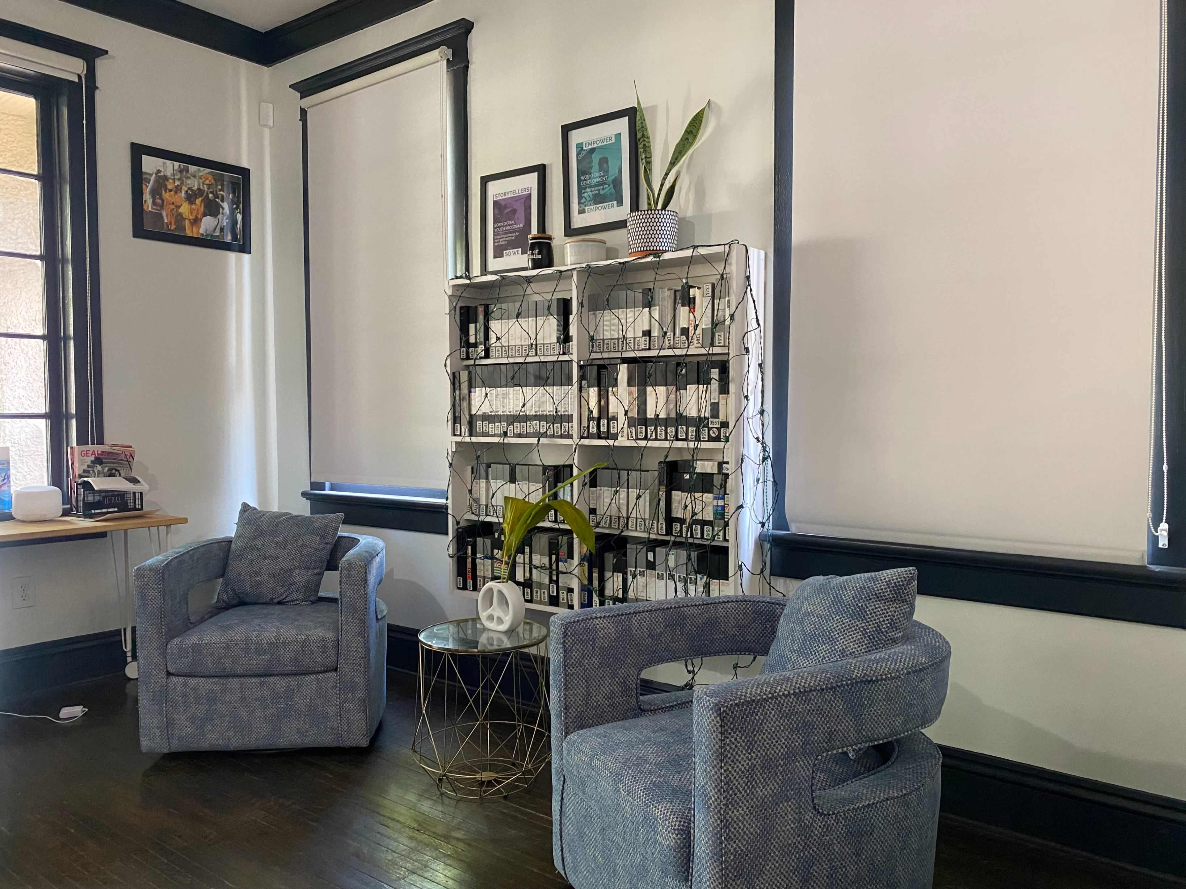 A cozy room with two blue armchairs positioned near a shelf filled with bottles and decorated with plants, alongside a small glass and metal side table.