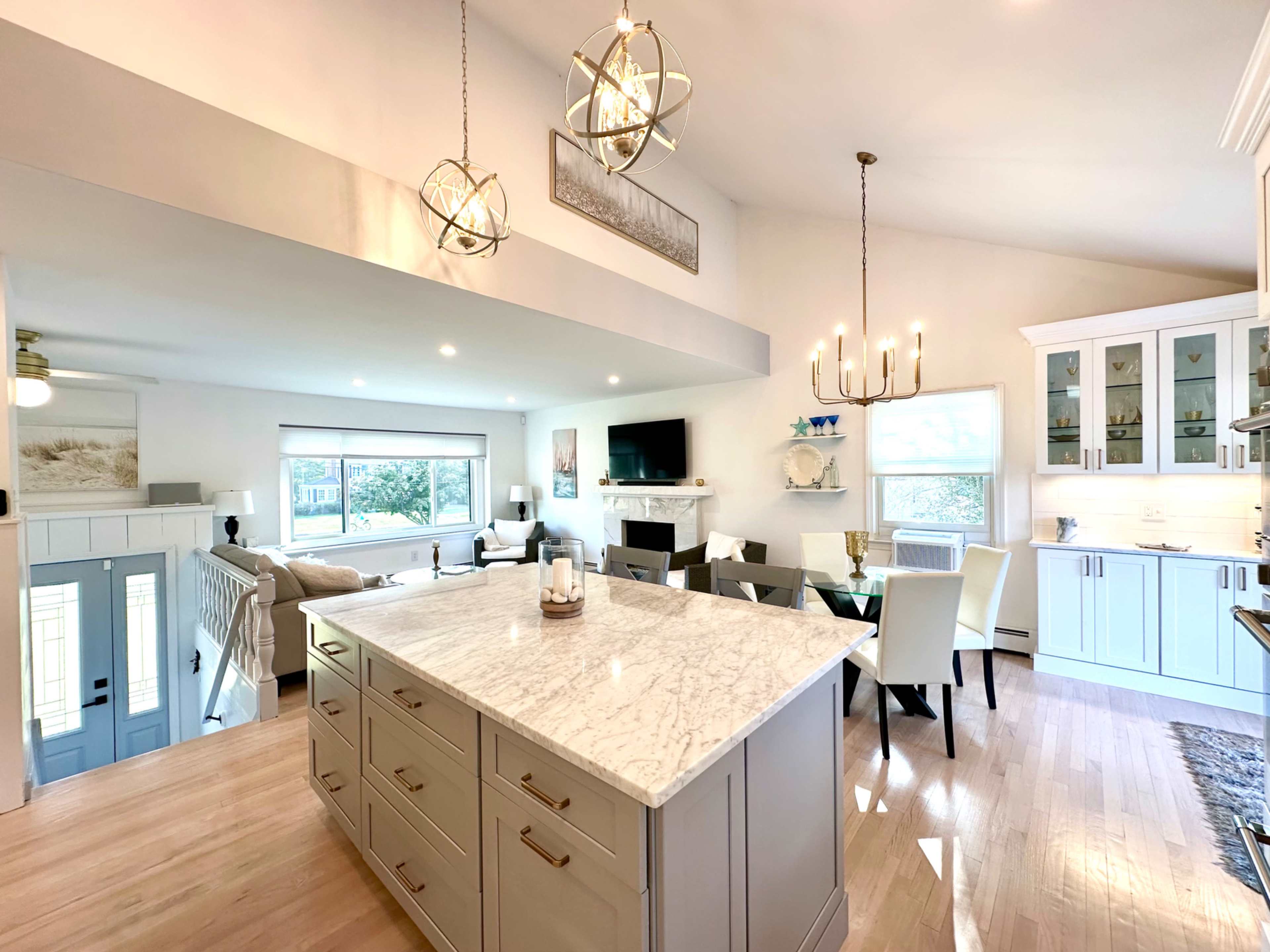 The image shows a bright, open-concept living space featuring a kitchen island, dining area, and a cozy seating area with large windows.