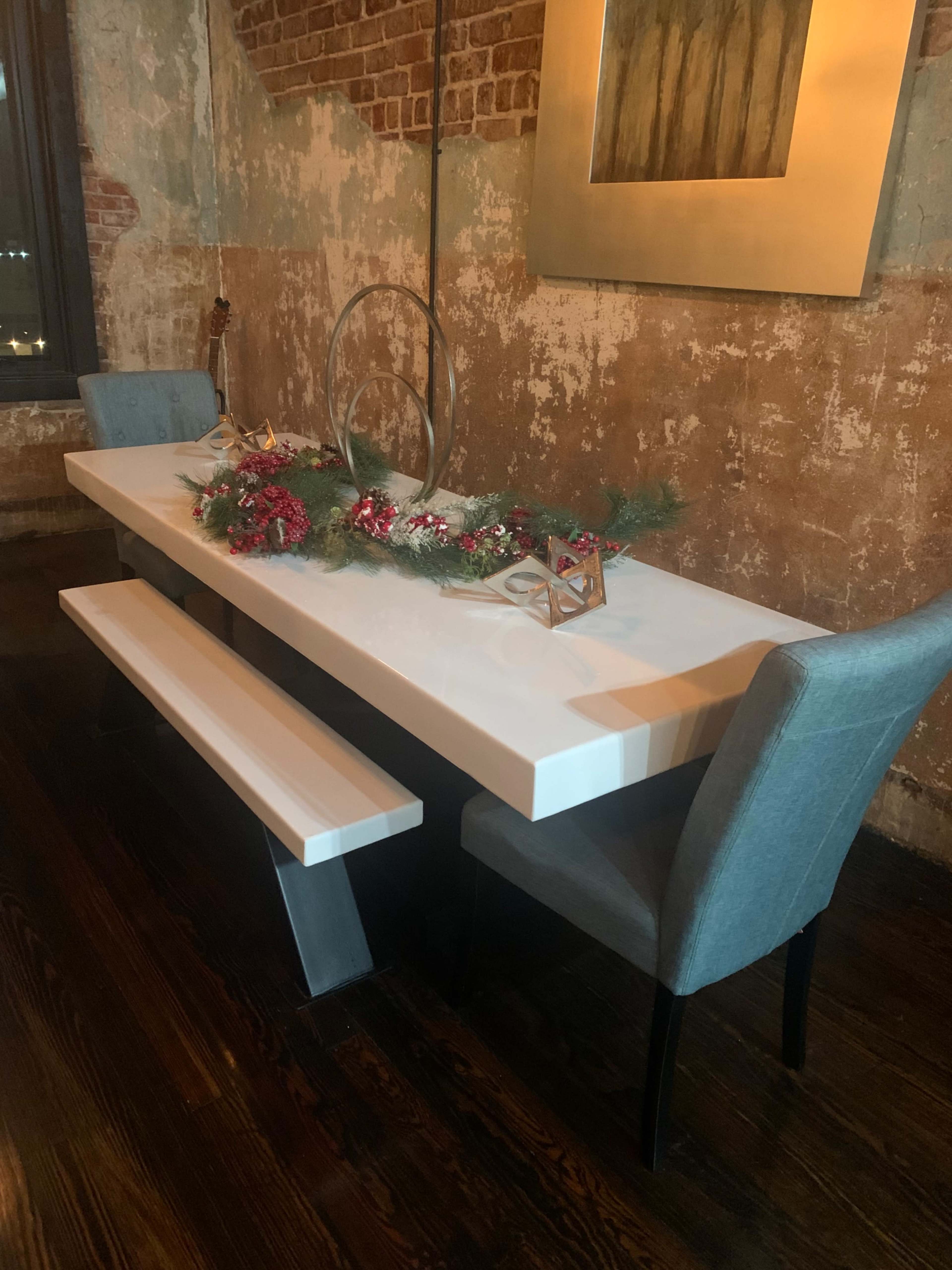 A modern dining table with a white surface is set against a rustic brick wall, adorned with greenery and decorations, accompanied by two gray upholstered chairs.