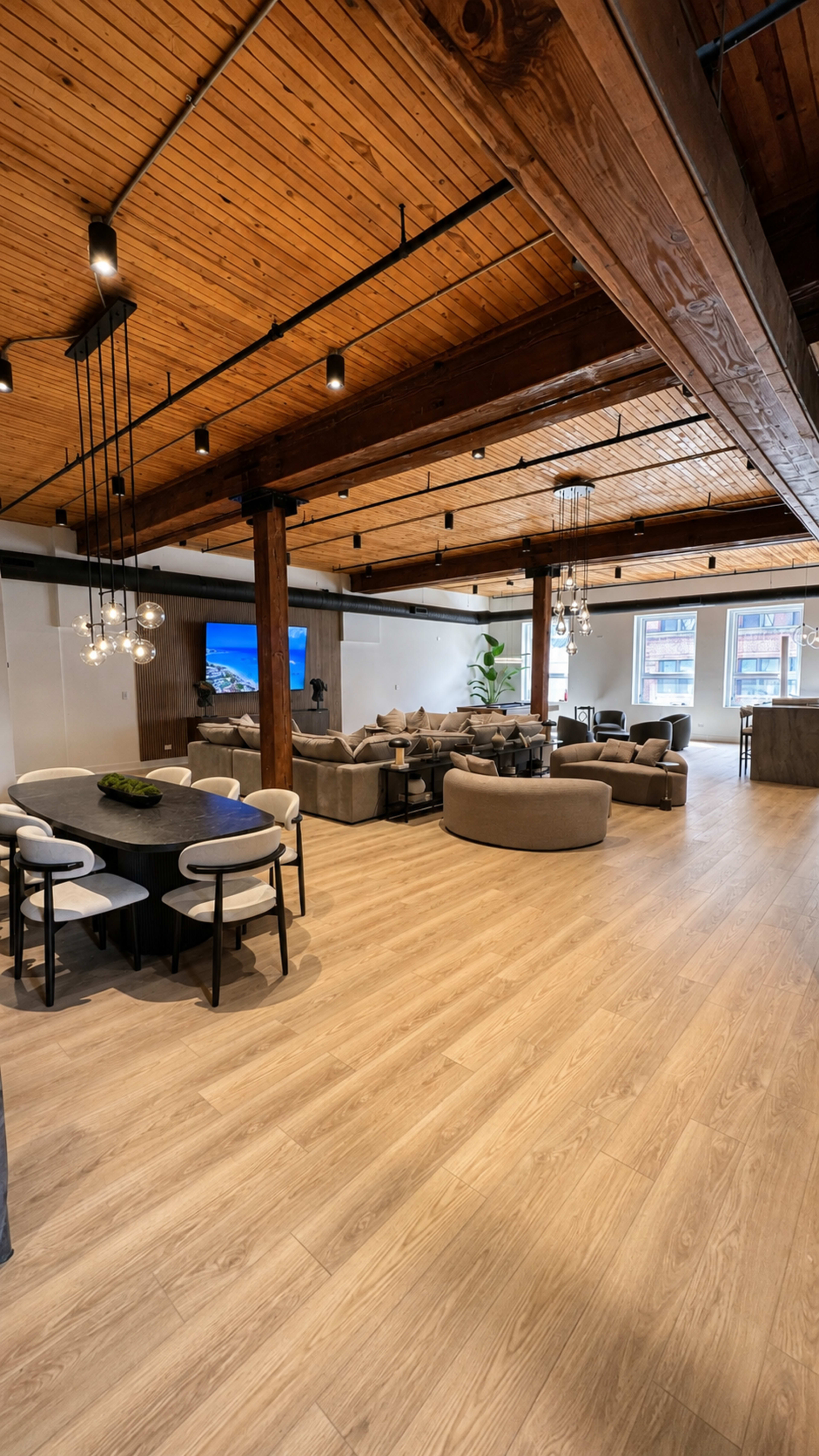 The image shows a spacious, modern interior featuring a dining area with a round table and four chairs on one side, and a cozy living area with multiple sectional sofas on the opposite side, all under wooden beams and soft lighting.