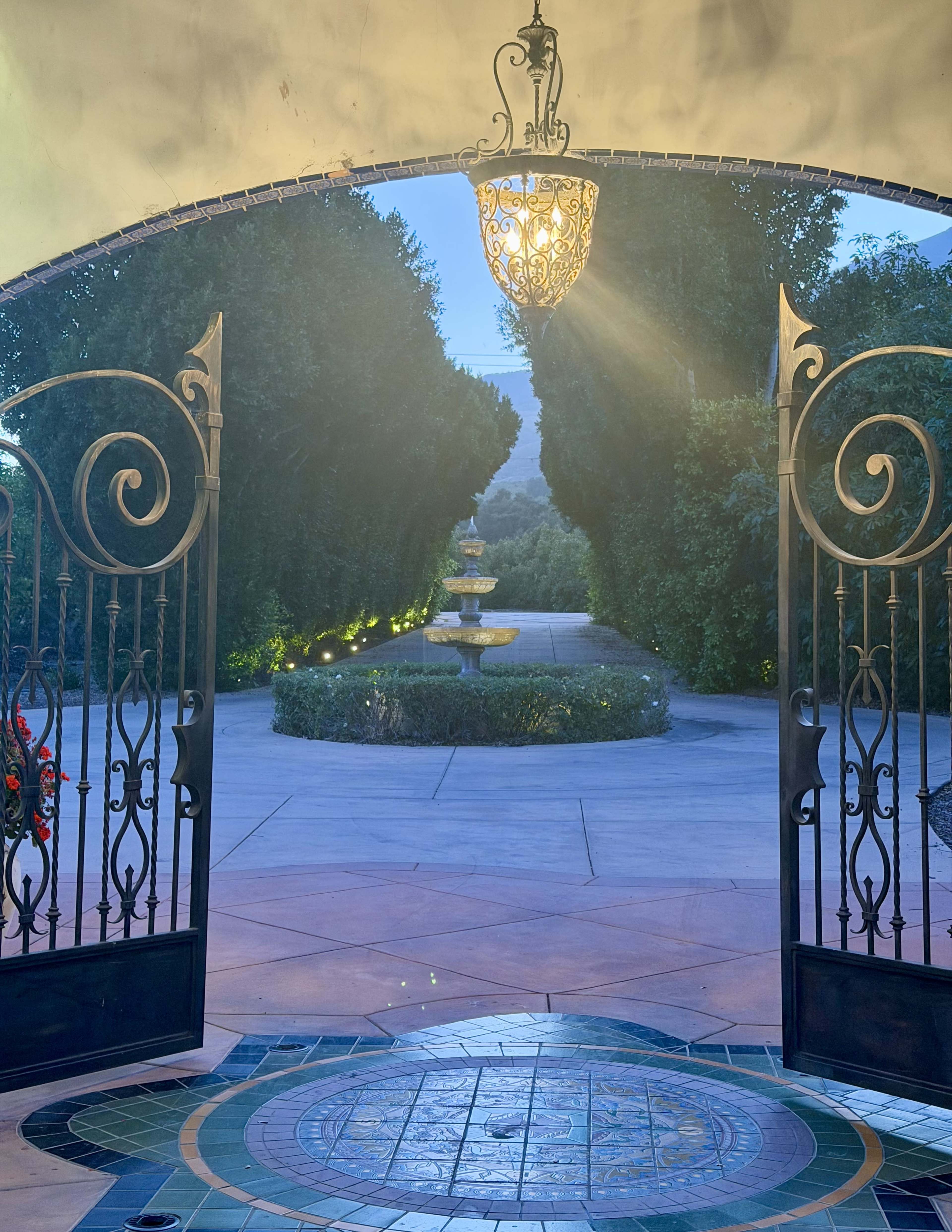 The image shows an ornate entrance gate opened towards a pathway featuring a fountain surrounded by trimmed hedges and soft lighting.