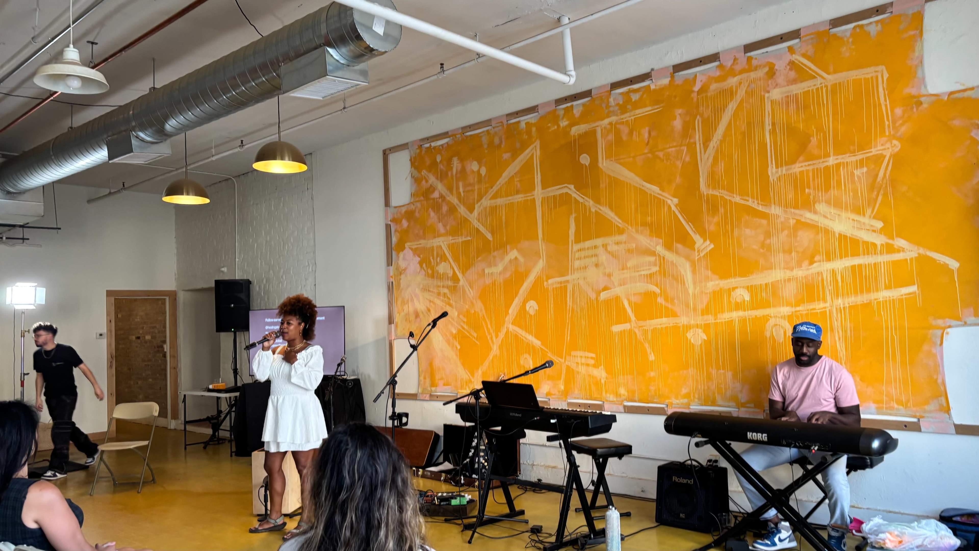 A performer in a white dress sings on stage while a musician plays the keyboard, and an audience observes in a bright, artistically decorated room with an orange mural on the wall.