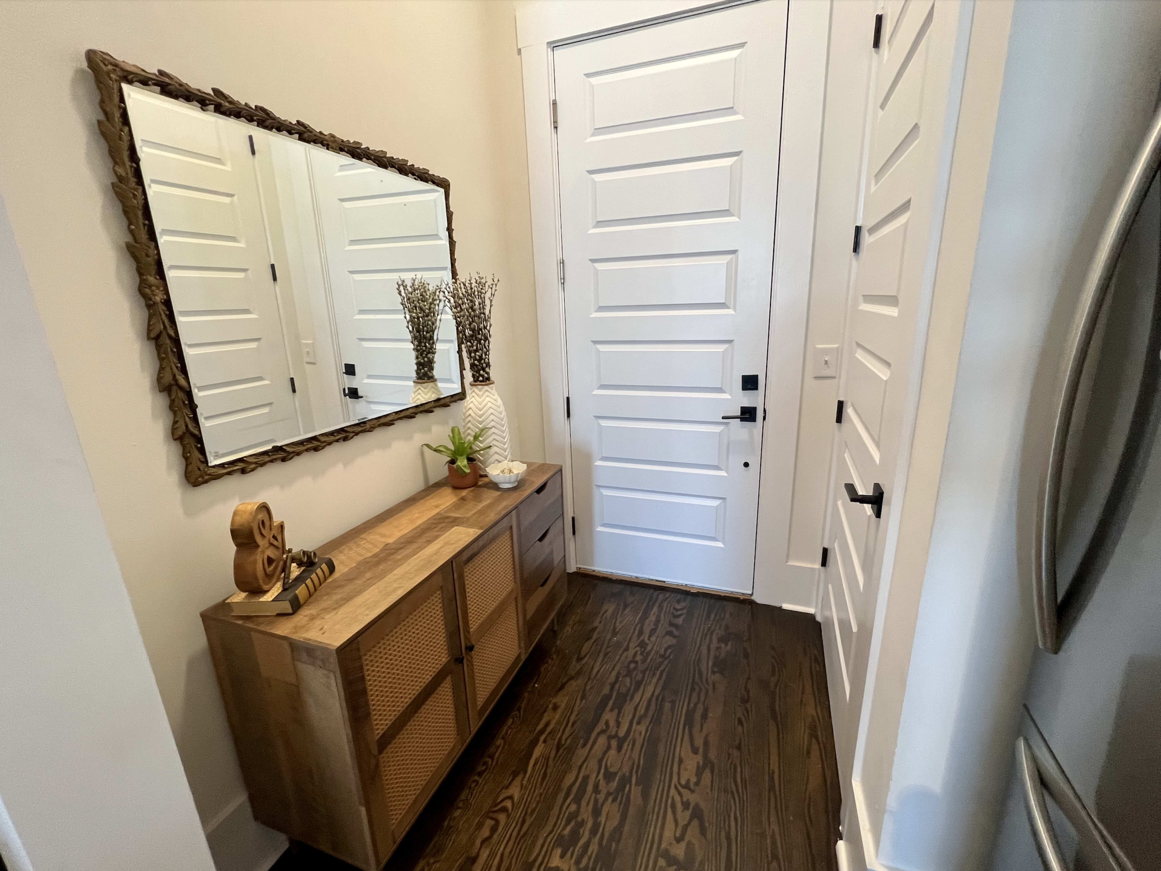 A hallway with a closed white door, a wooden console table with decorative items, a large mirror, and a small plant.