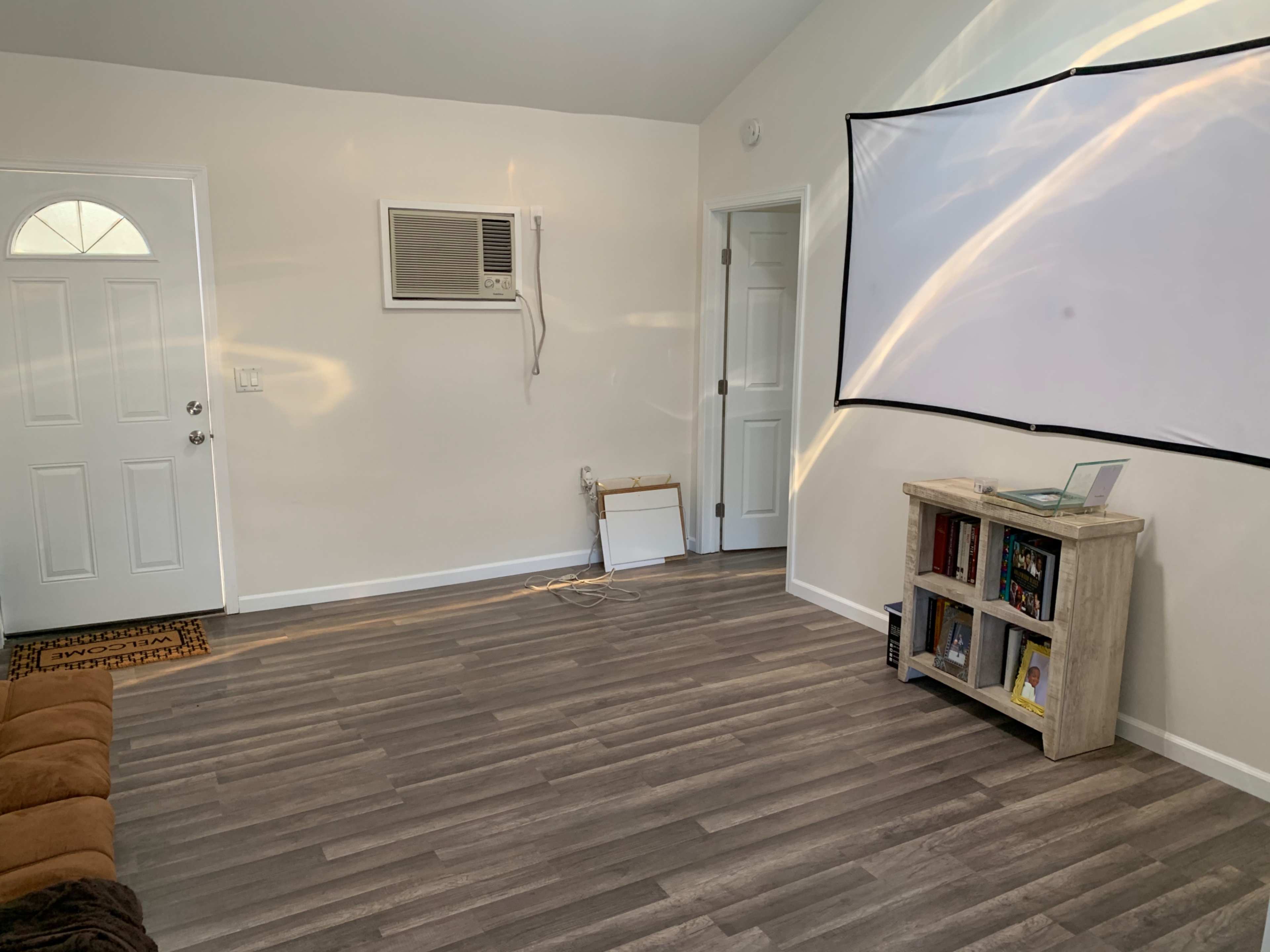 The image shows a small room with a door, an air conditioning unit, a white wall-mounted screen, and a bookshelf against one wall.