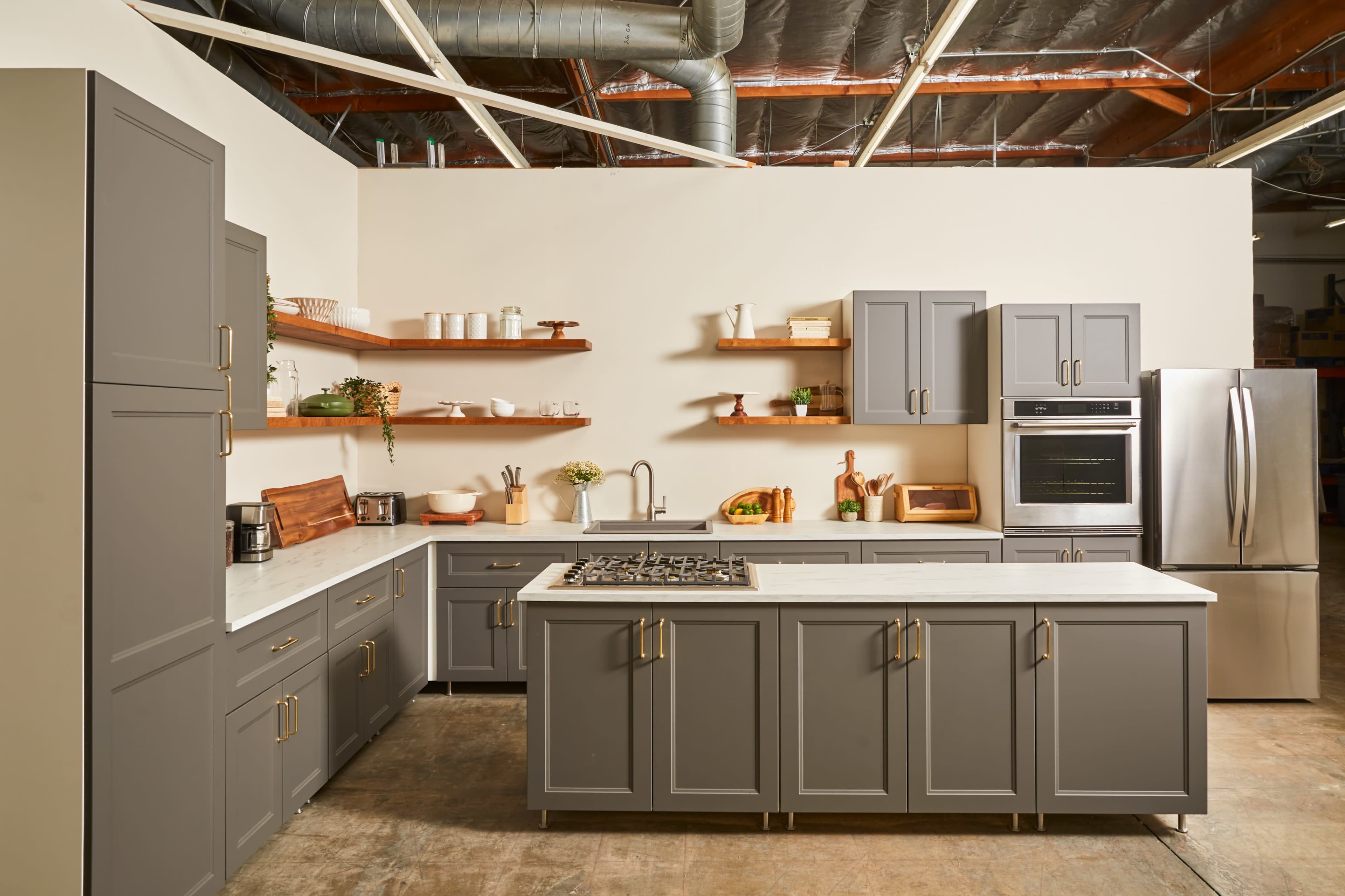 A modern kitchen with gray cabinets, a large island with a cooktop, open shelving, and stainless steel appliances.