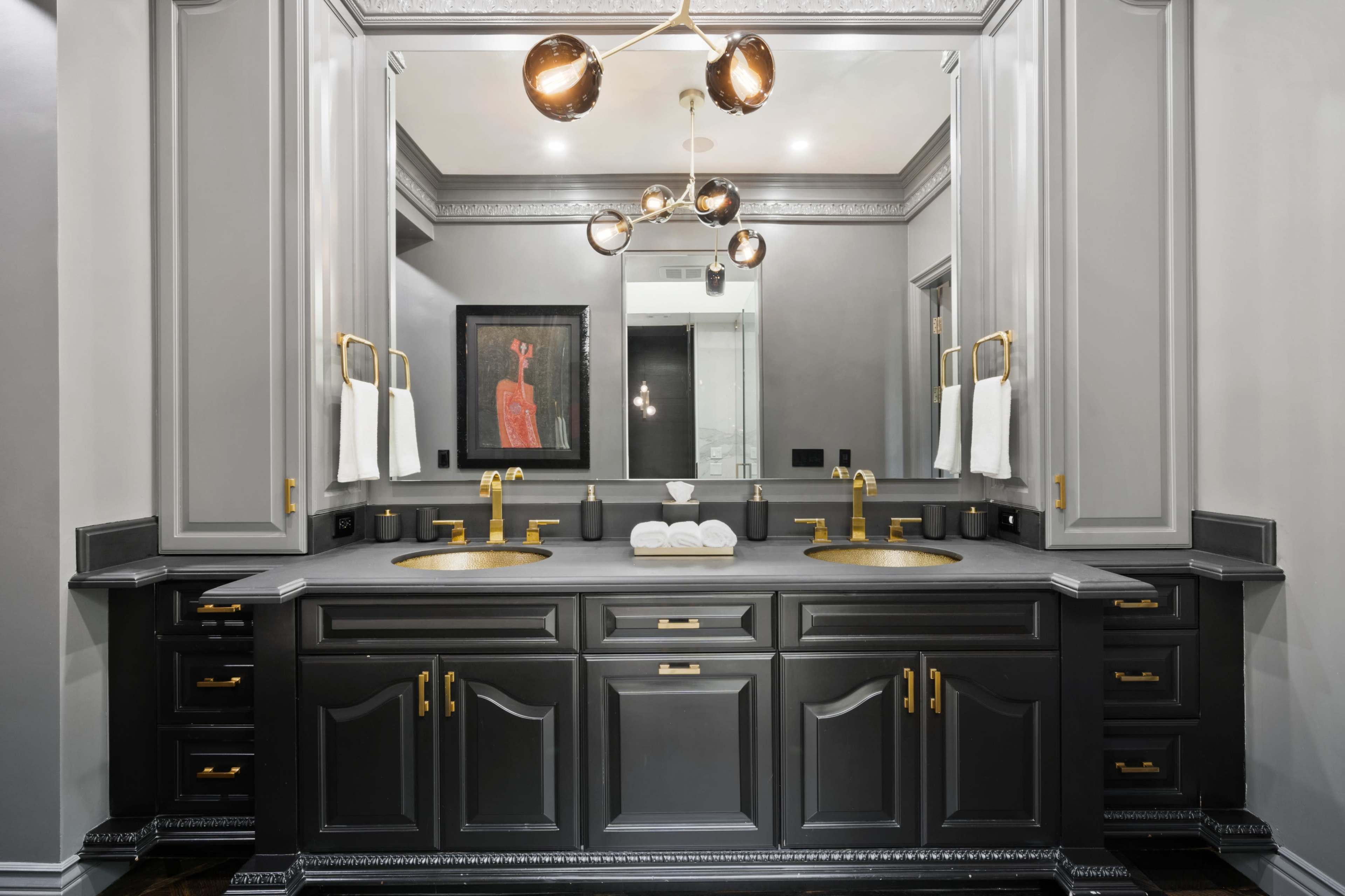 The image shows a modern bathroom vanity with dual sinks, a dark cabinetry finish, and gold fixtures, framed by large mirrors.