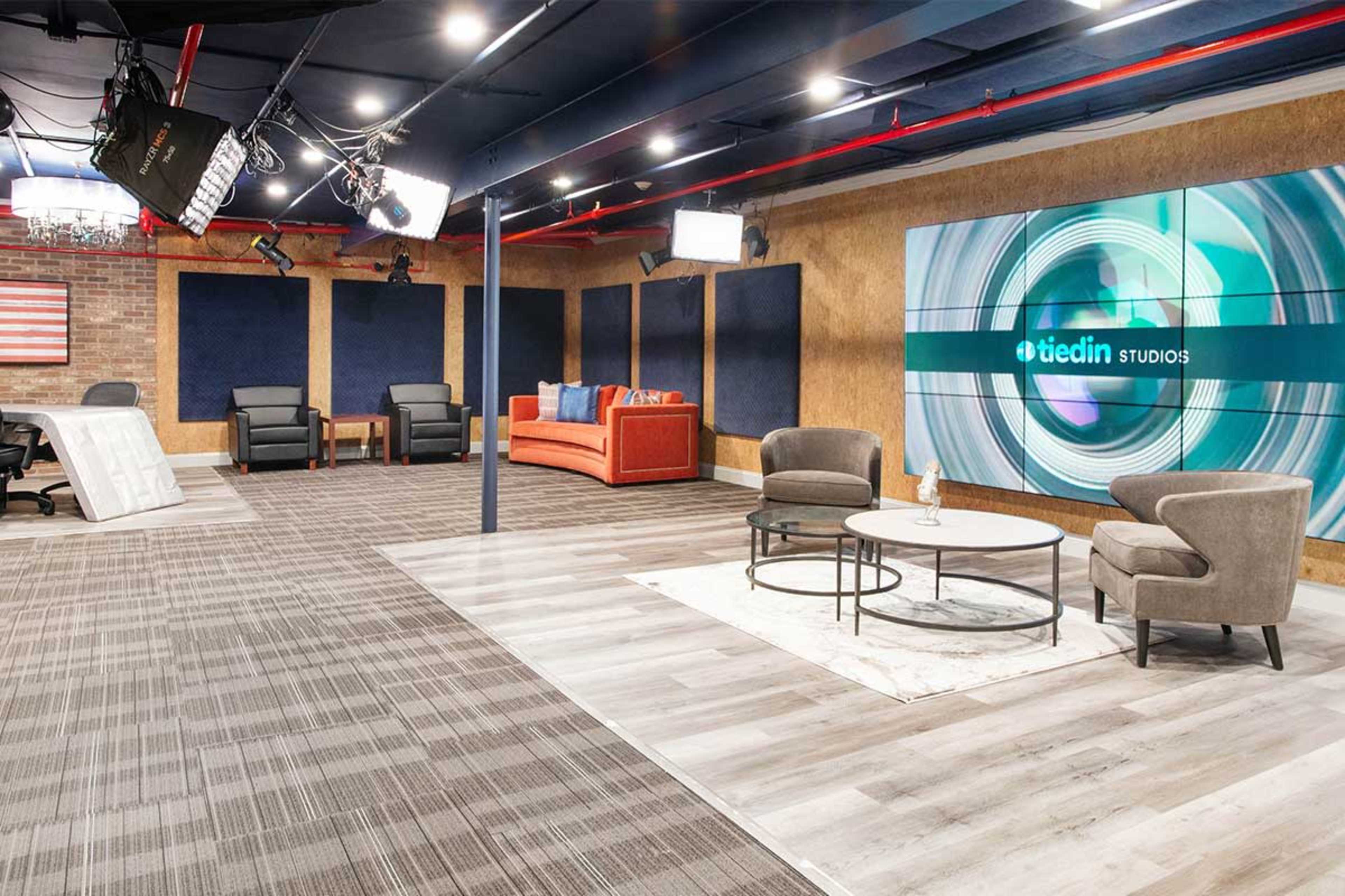 The image depicts a modern studio setting featuring a beige and blue color scheme, with comfortable seating, professional lighting, and a large screen displaying the logo of a media company.