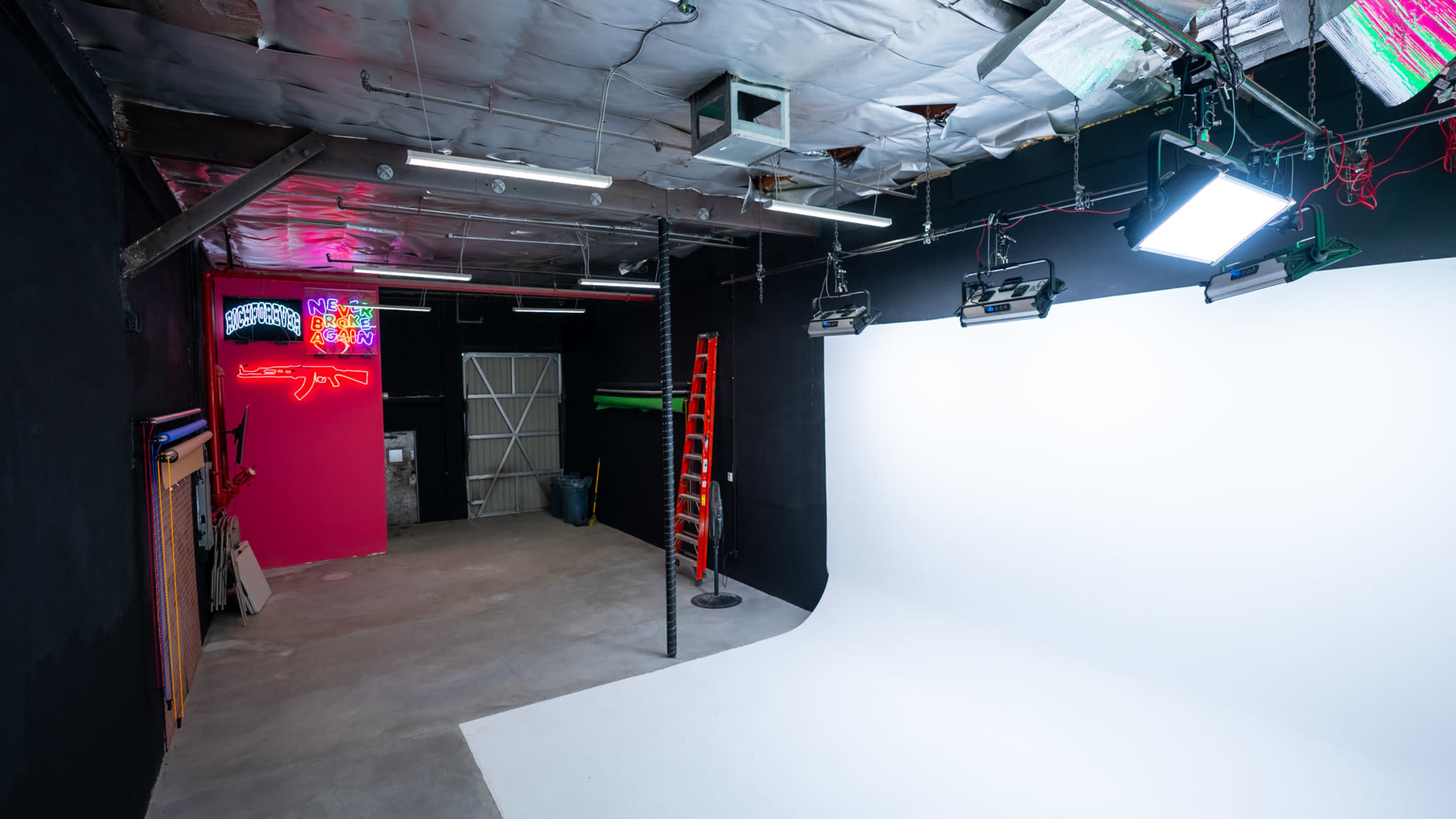 A photography studio with a white backdrop, lighting equipment, and a ladder, set against black walls and a pink accent wall.