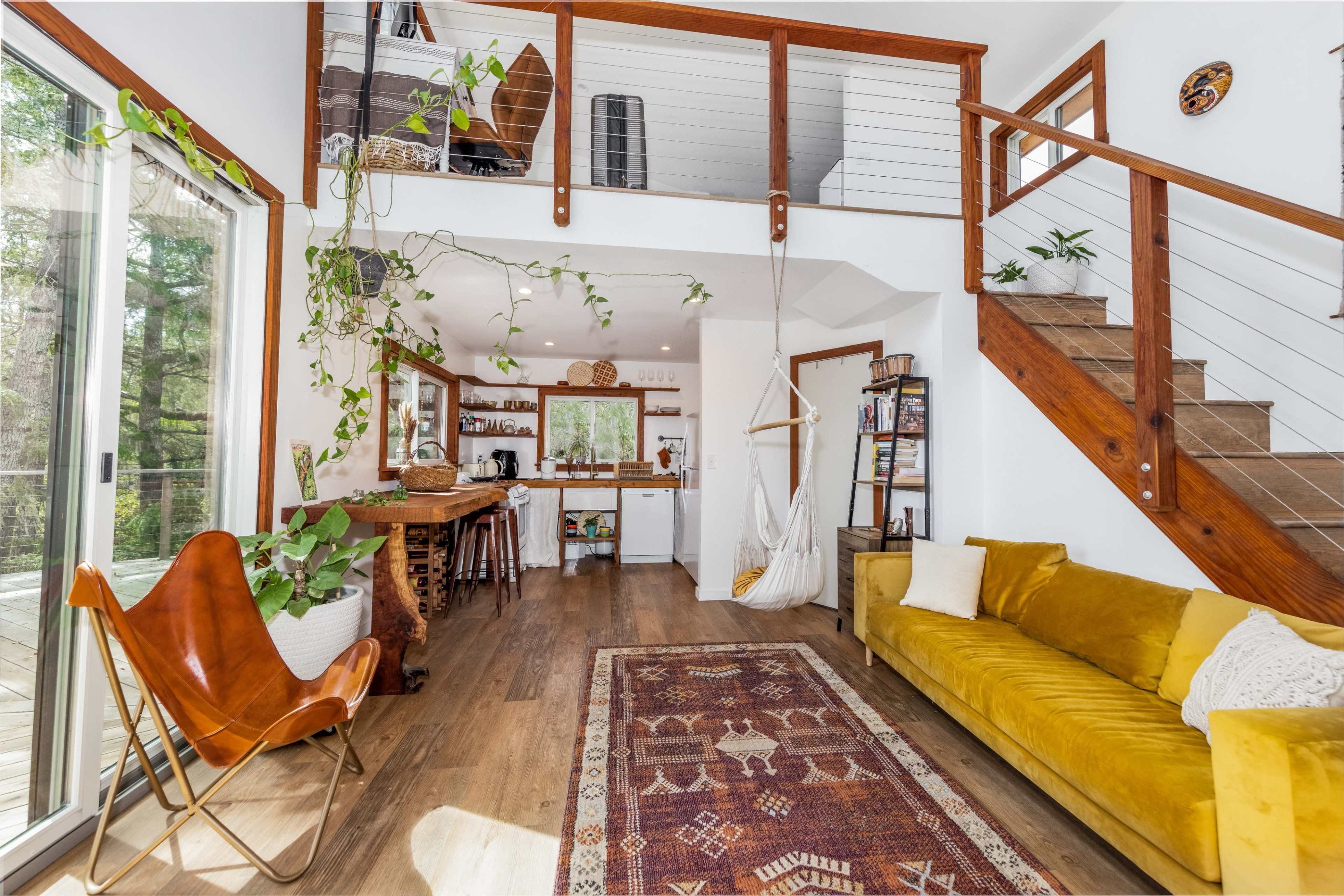 A cozy living area with wooden stairs leading to an upper level, a yellow couch, a hanging chair, and plants adorning the space, alongside a kitchen in the background.