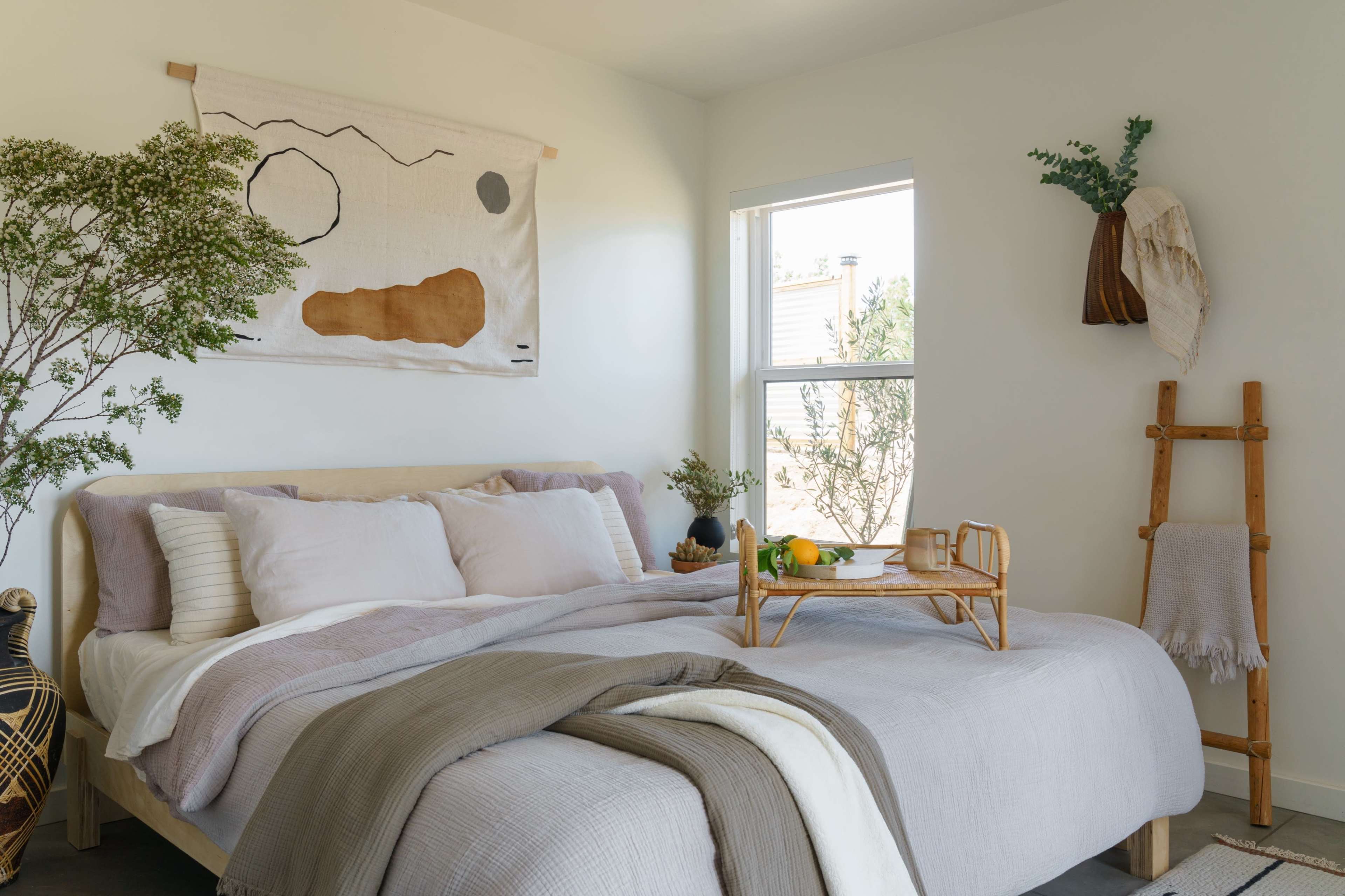 A neatly arranged bedroom features a queen-sized bed with layered bedding, a wooden tray with fruits and drinks, a wall tapestry, and a window allowing natural light in.
