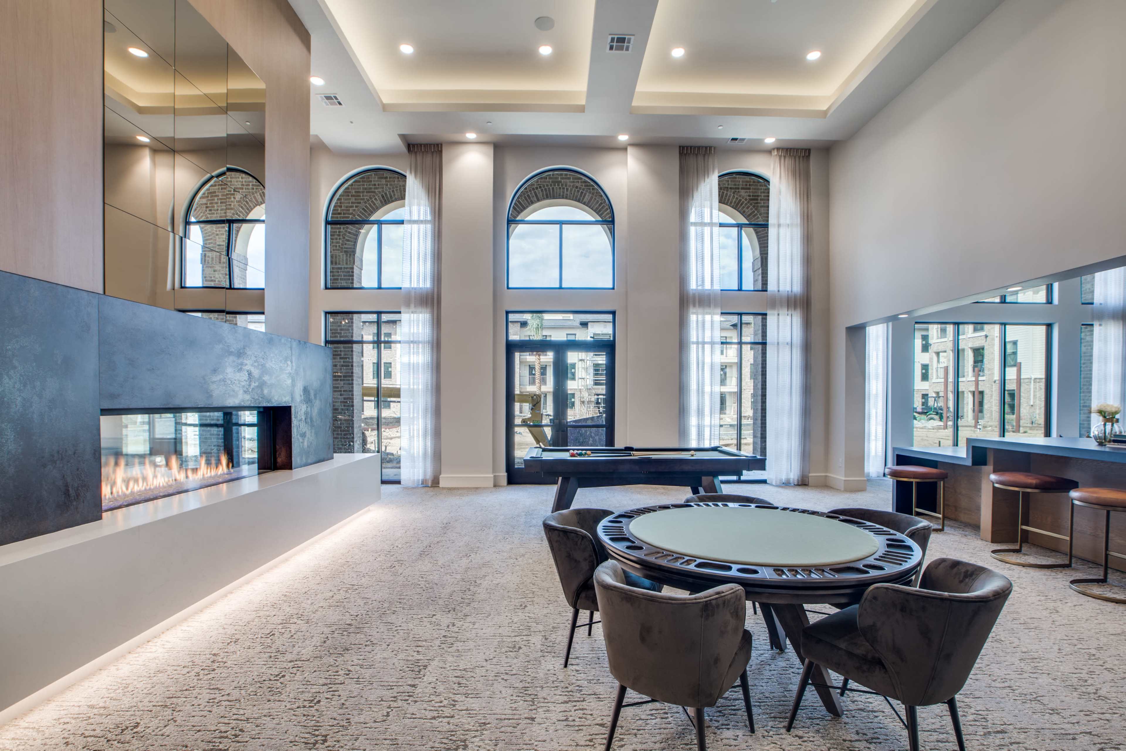The image shows a modern lounge area with large windows, a fire feature, a poker table, and a pool table in a well-lit space.