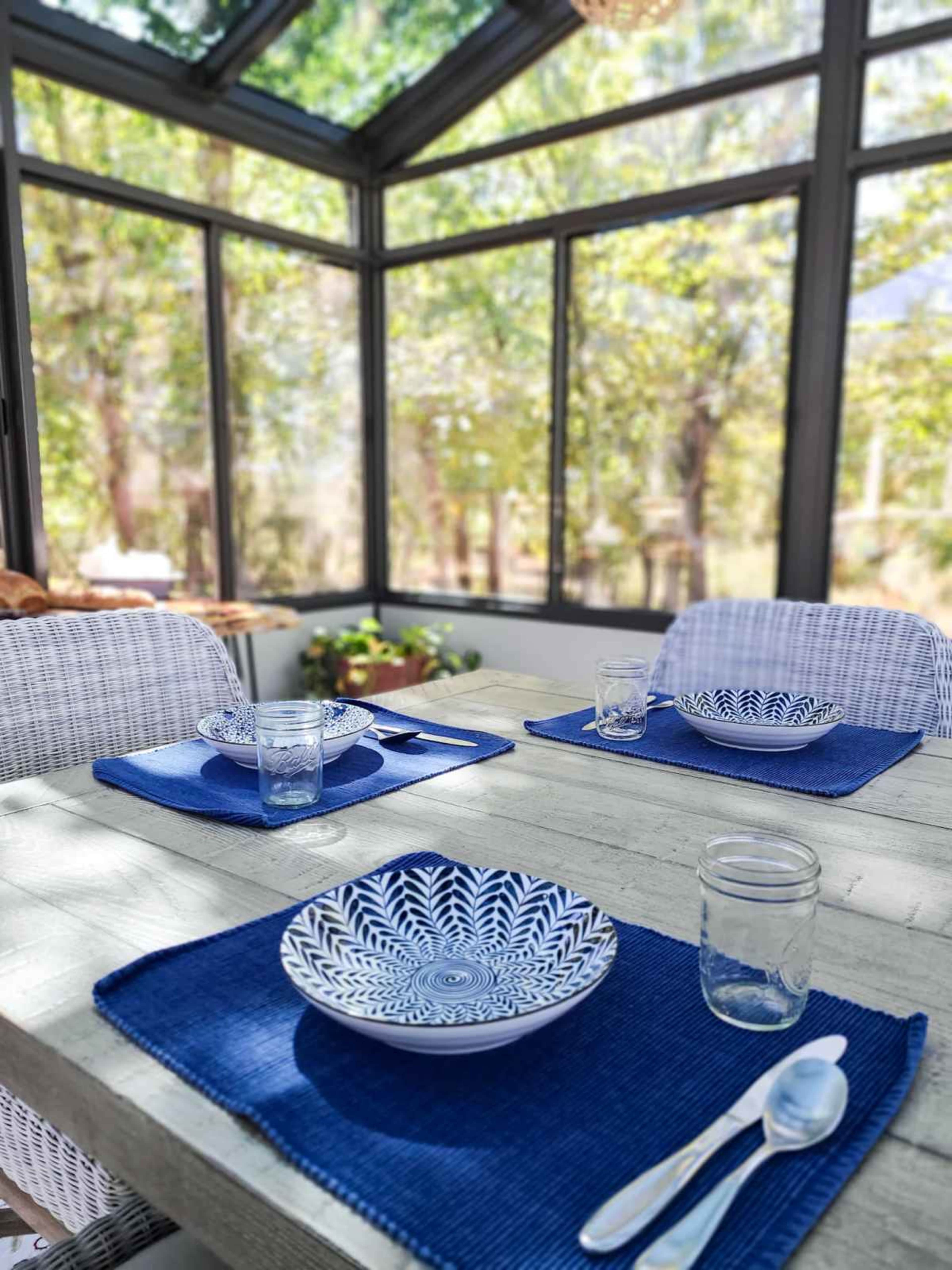A dining table is set with patterned plates and glassware in a sunlit, glass-enclosed room surrounded by trees.
