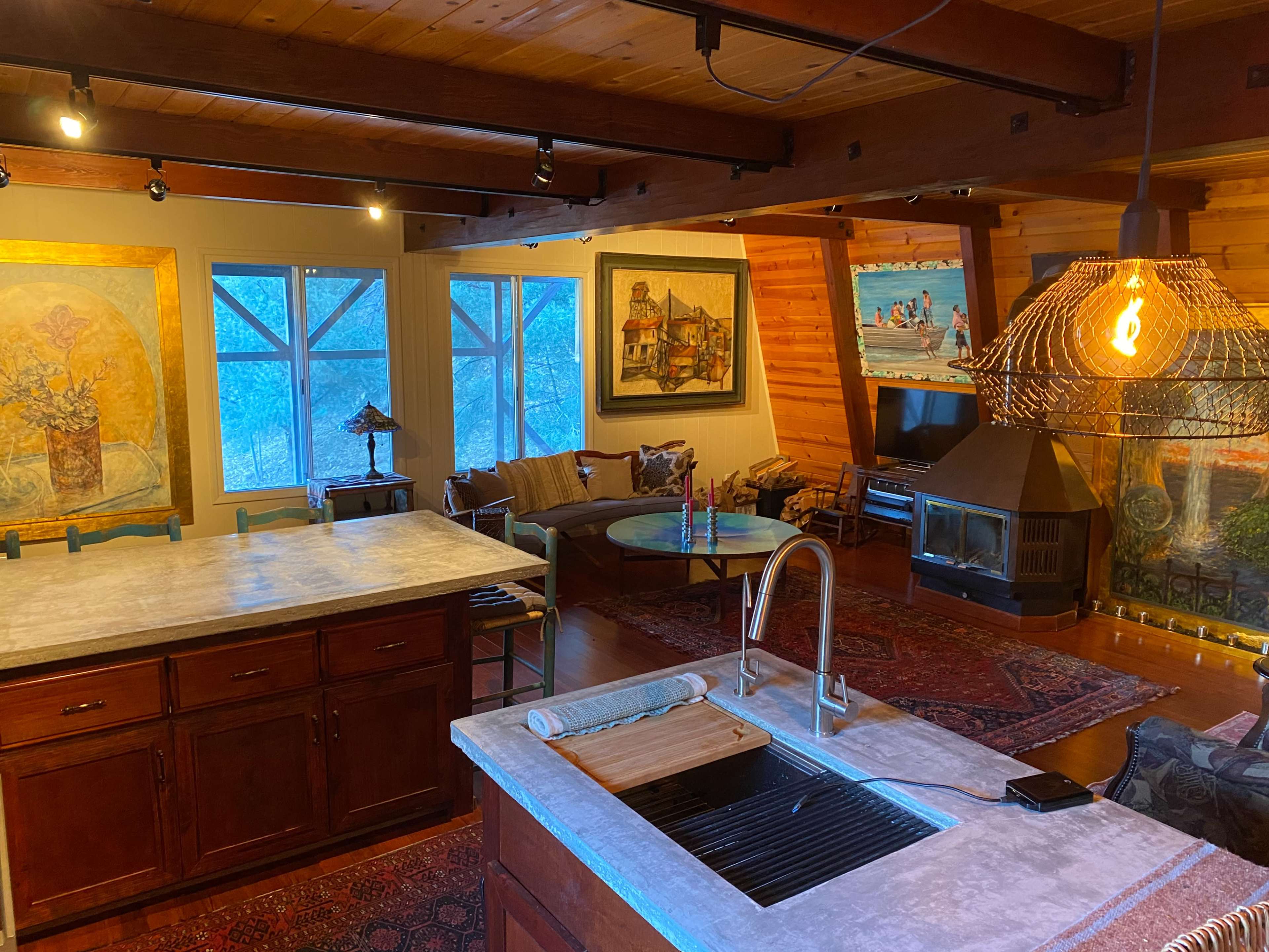 The interior of a cozy, well-lit living space features a kitchen island, a seating area with large windows, and several colorful paintings on the walls.