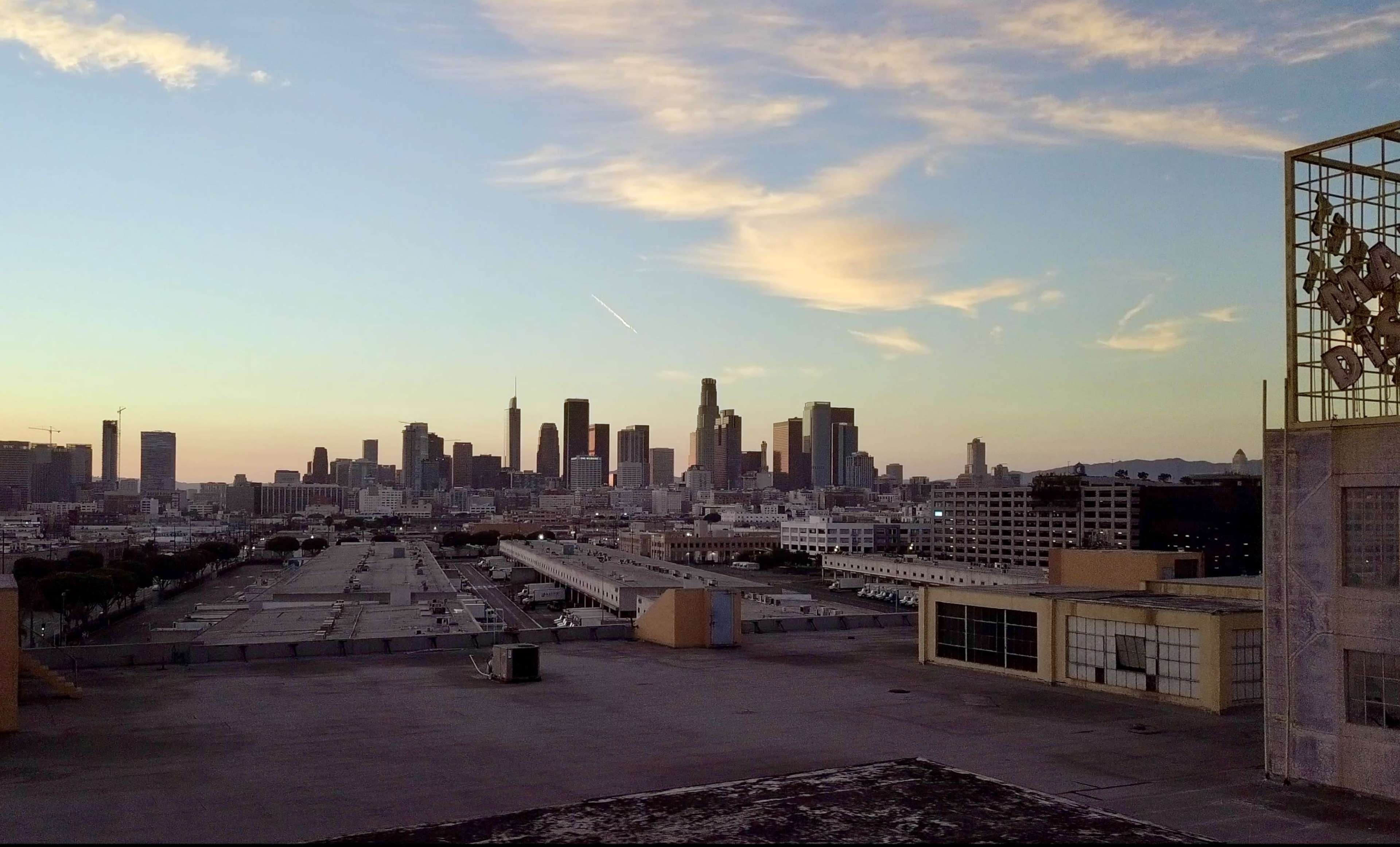 A view of the Los Angeles skyline at sunset, with buildings silhouetted against the colorful sky and rooftops in the foreground.